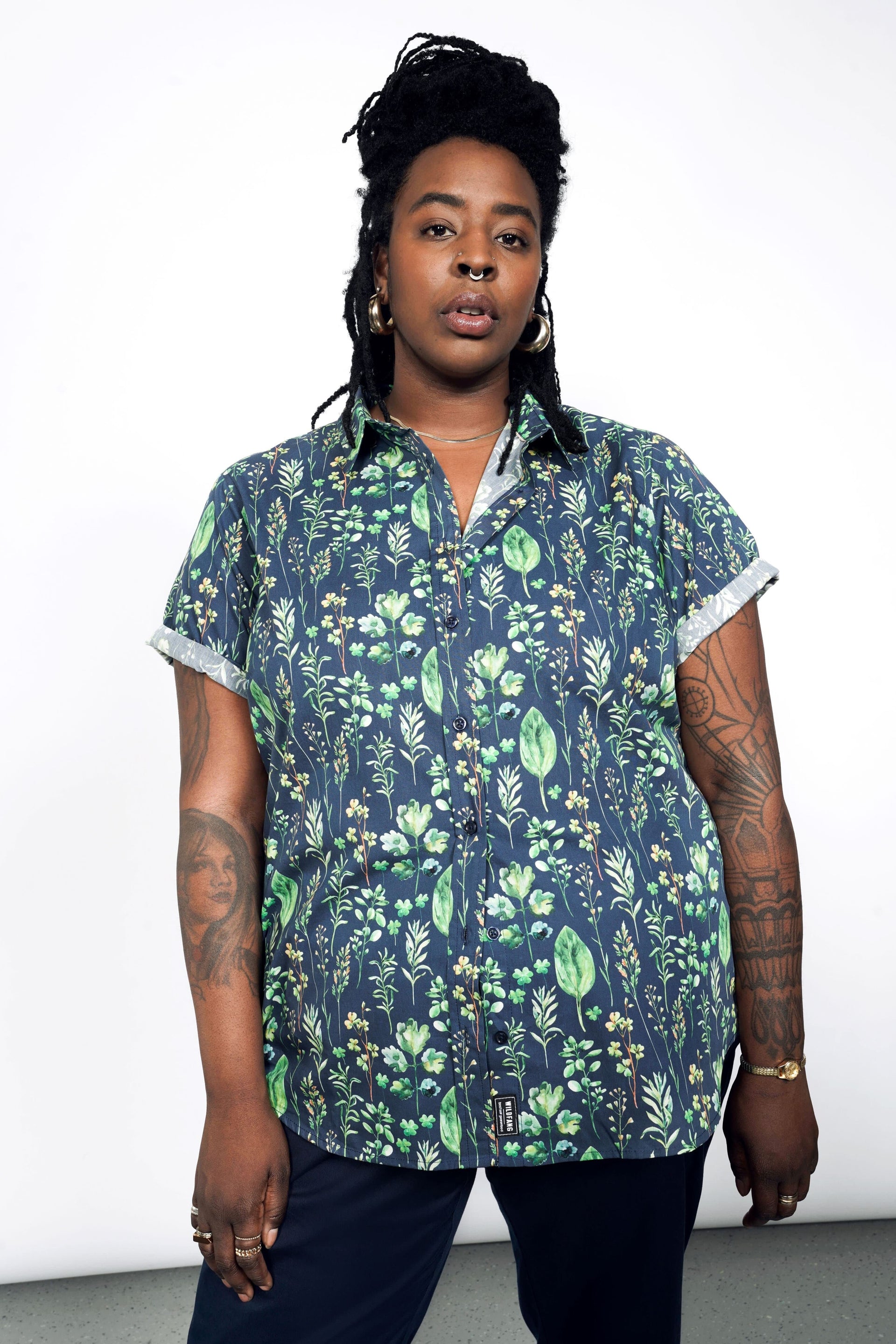 Wearing The Essential Button Up, a tailored, navy blue shirt with green floral patterns and short sleeves, made of 100% cotton, they stand confidently against a plain backdrop. Dreadlocks frame their face, and tattoos are visible on their arms as they gaze at the camera.