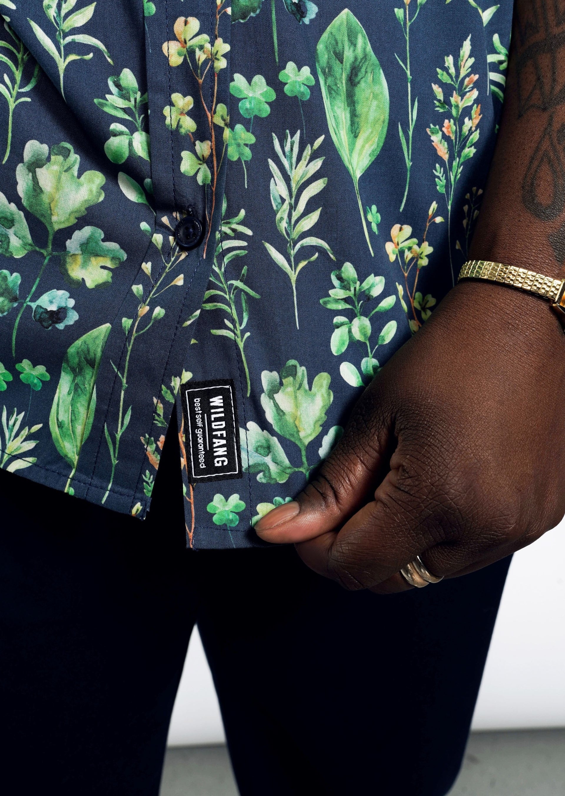 A person wearing The Essential Button Up, a tailored fit, short-sleeved, 100% cotton shirt with a dark floral pattern and visible WILDFANG label, holds the hem. Their dark skin is accented by a gold ring and bracelet.