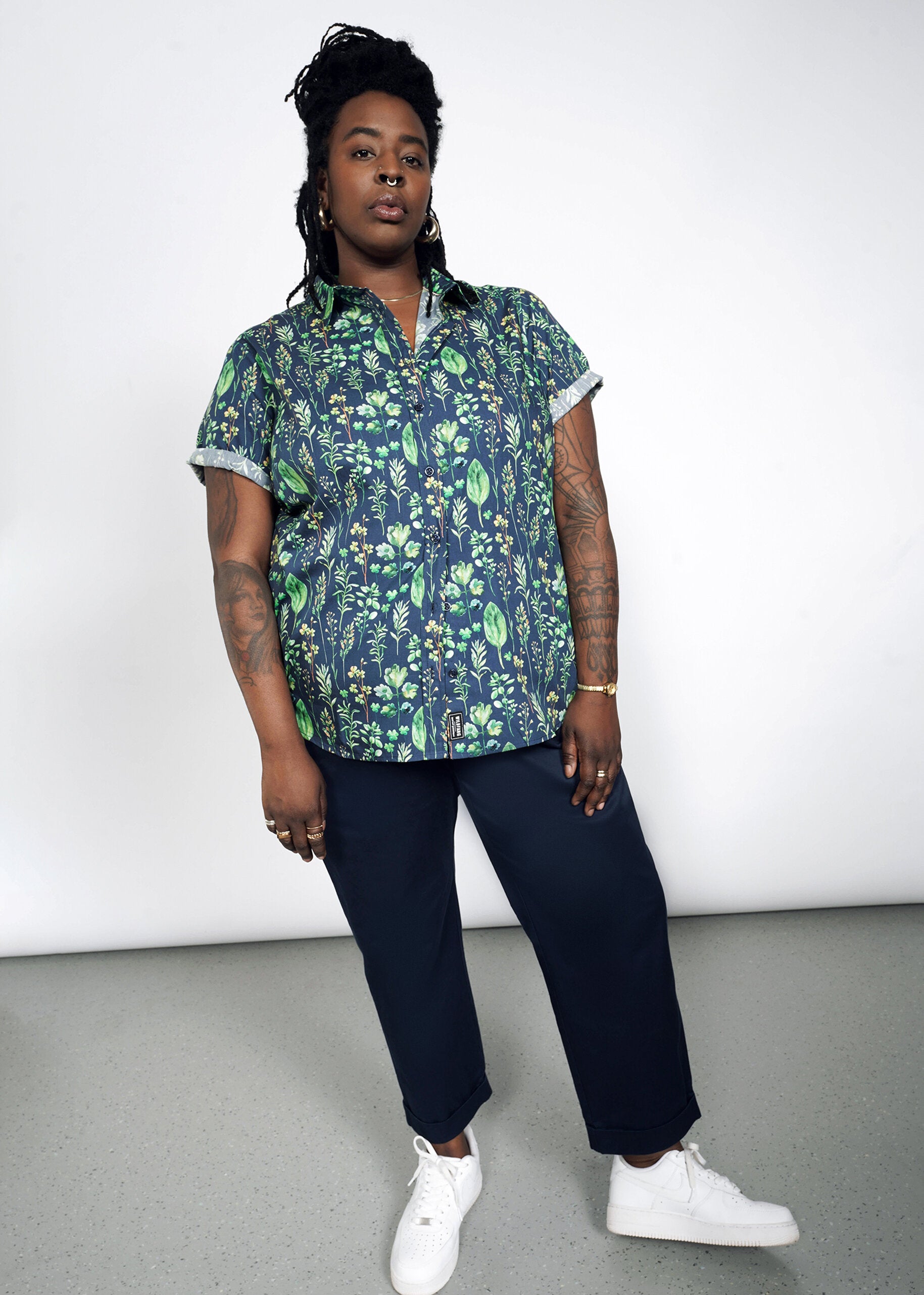 A person with long hair and tattoos confidently wears The Essential Button Up, a tailored fit, short-sleeved green and blue floral shirt made from 100% cotton, paired with dark pants and white sneakers. The background is plain and light-colored.