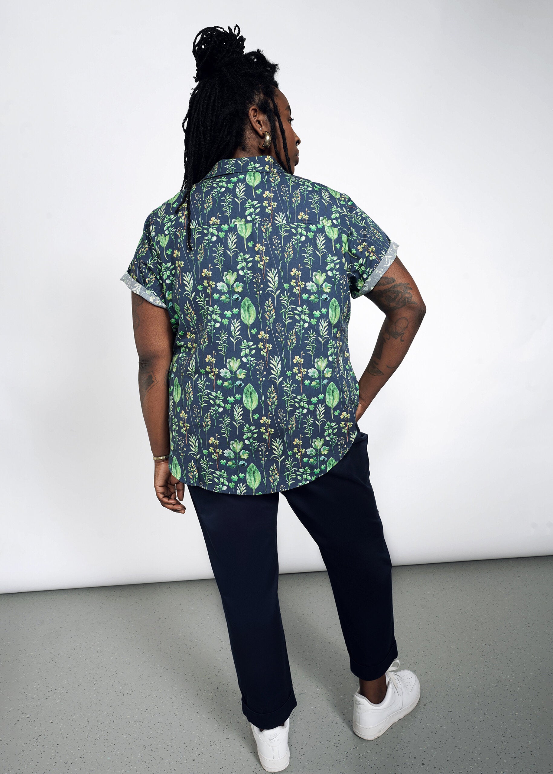 Wearing The Essential Button Up, a short-sleeved, 100% cotton shirt in a tailored fit with a green leaf pattern, the person with long braided hair stands back to the camera. They pair it with dark pants and white sneakers against a plain, minimalistic background.