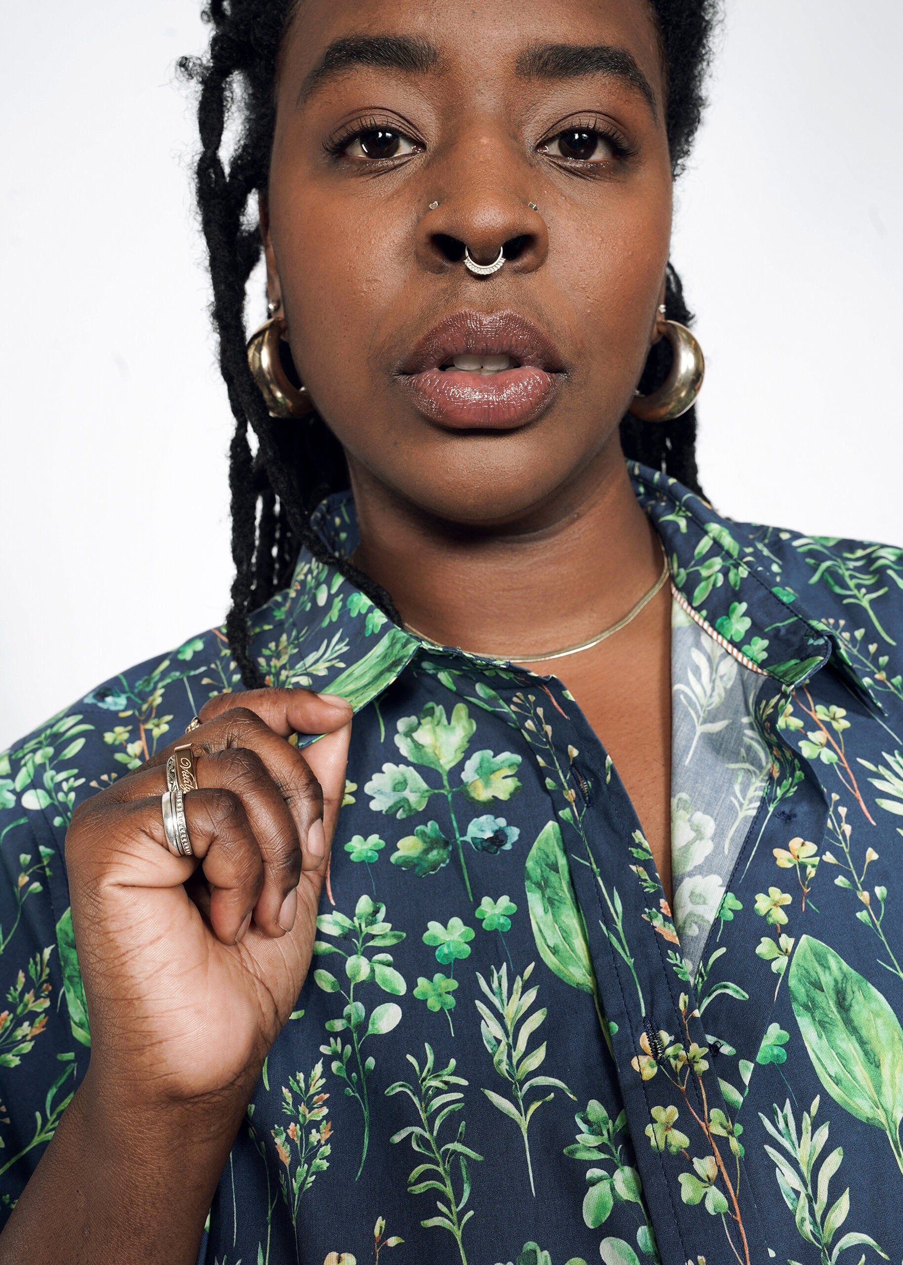 A person with long braided hair, hoop earrings, and a nose ring is wearing The Essential Button Up—a tailored short-sleeve dark floral-patterned shirt made of 100% cotton. They are holding the collar against a plain white background.