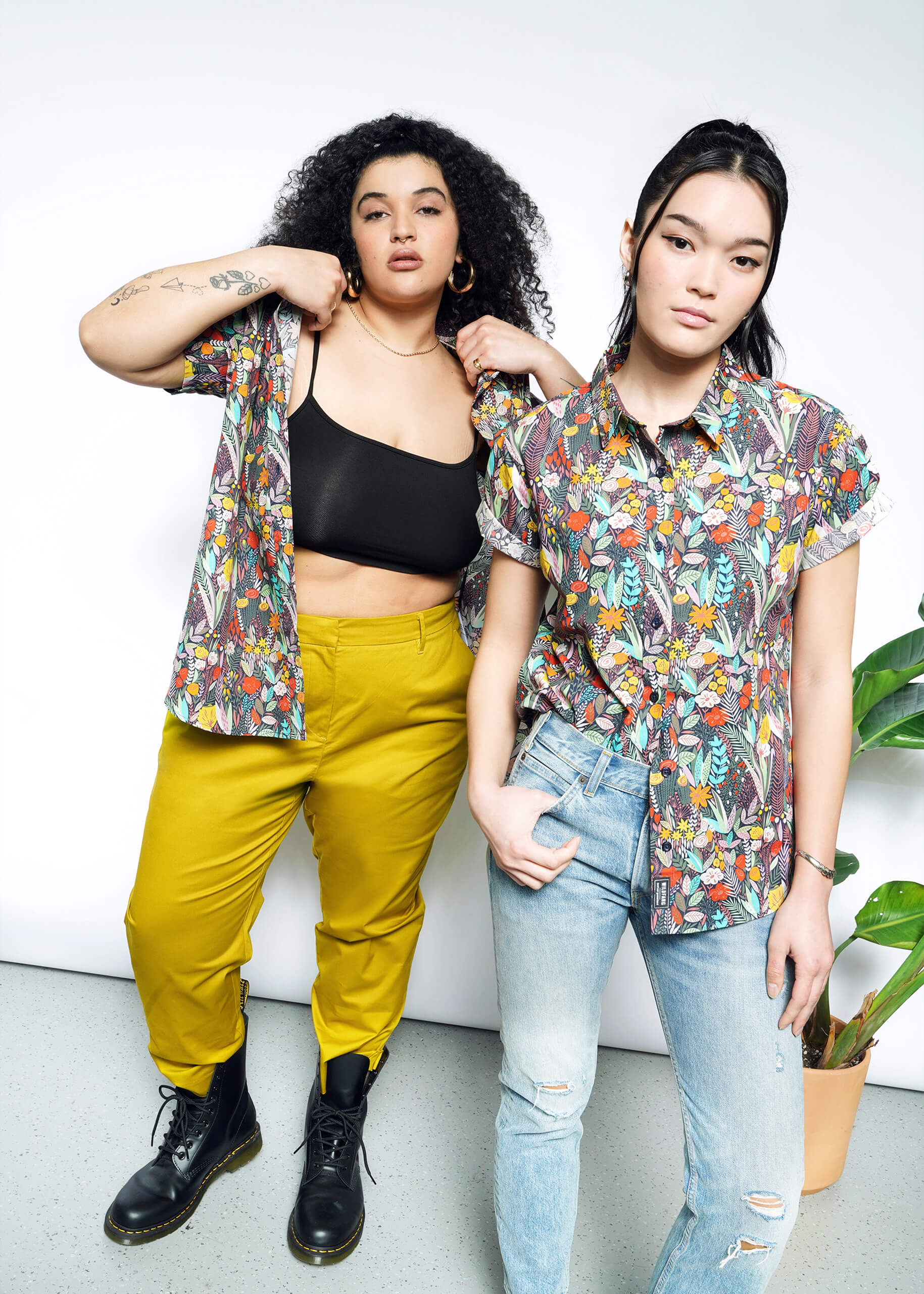 Two people stand in a bright room wearing colorful shirts. The person on the left sports yellow pants and boots, while the one on the right pairs ripped jeans with sneakers. The vibrant Essential Button Up adds flair, complemented by breathable cotton fabric. A plant is visible on the side.