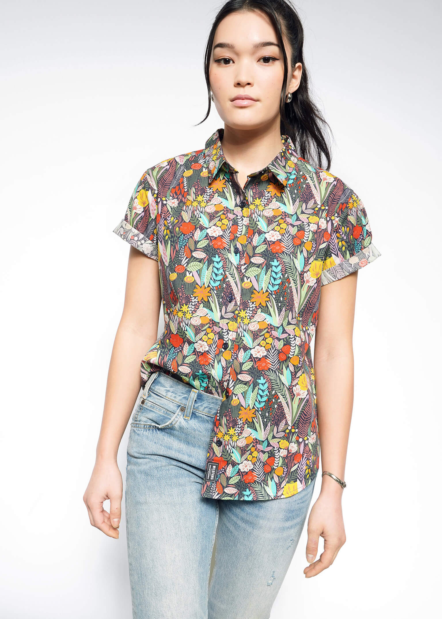 A person wearing The Essential Button Up, a short-sleeved floral shirt in breathable cotton, pairs it with light blue jeans. Their hair is tied back, and they maintain a neutral expression against a plain white background.