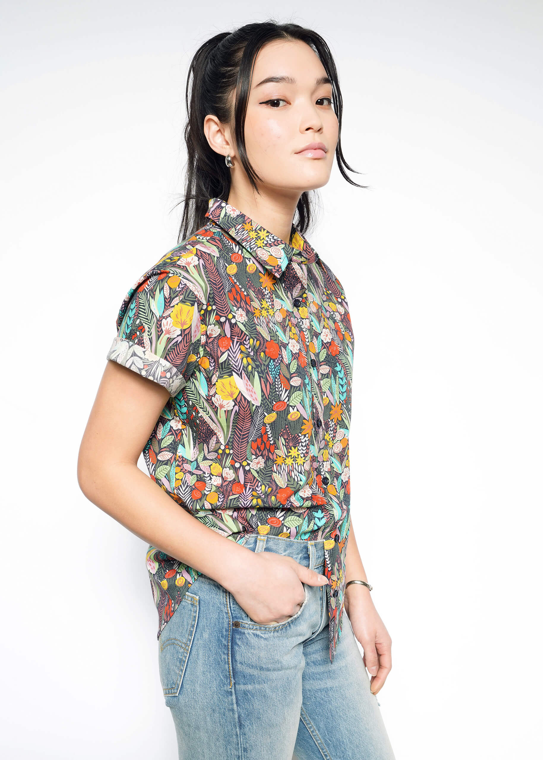 A person stands against a light background wearing The Essential Button Up, a colorful patterned short-sleeve shirt in breathable cotton, paired with blue jeans. Their hair is styled in a ponytail with loose strands framing their face, one hand in a pocket, and a neutral expression.