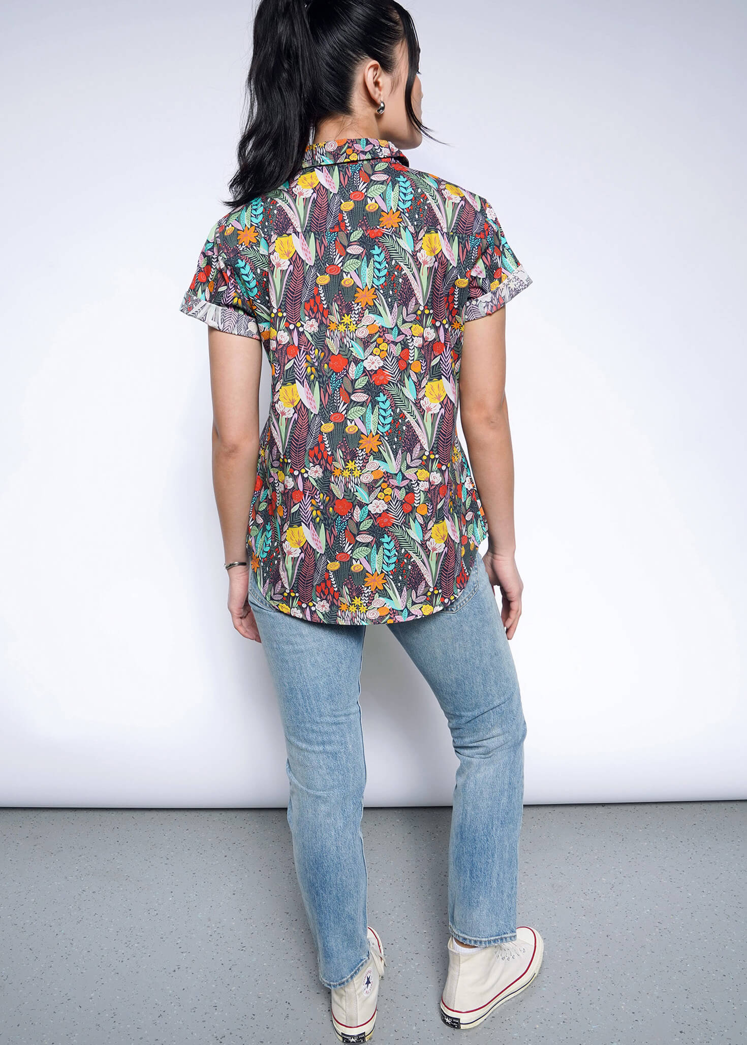 A person with long dark hair in a ponytail wears The Essential Button Up, a colorful floral short-sleeved shirt made from breathable cotton, paired with light blue jeans and white sneakers. They face away from the camera against a light background.