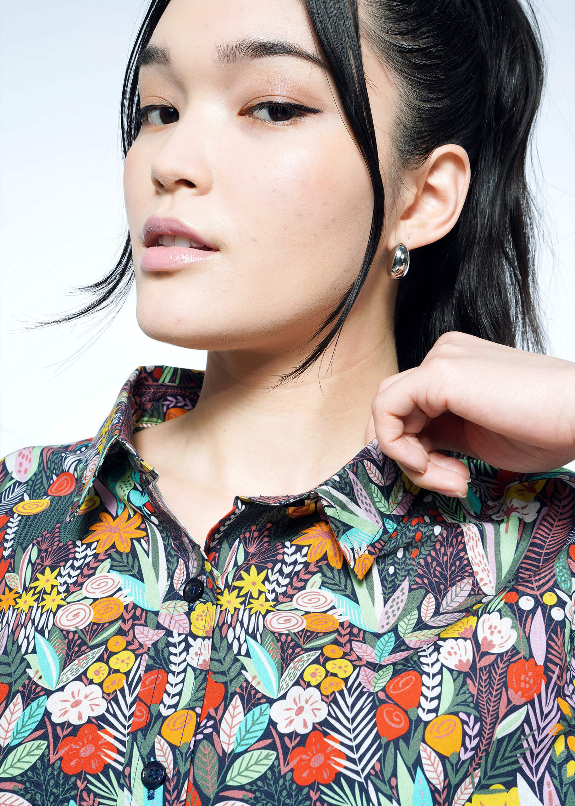 Close-up of a person with dark hair tied back, wearing The Essential Button Up by Wildfang, featuring vibrant leaf and flower patterns. Made from breathable cotton for a tailored fit, they have a neutral expression while holding the shirts collar with one hand.