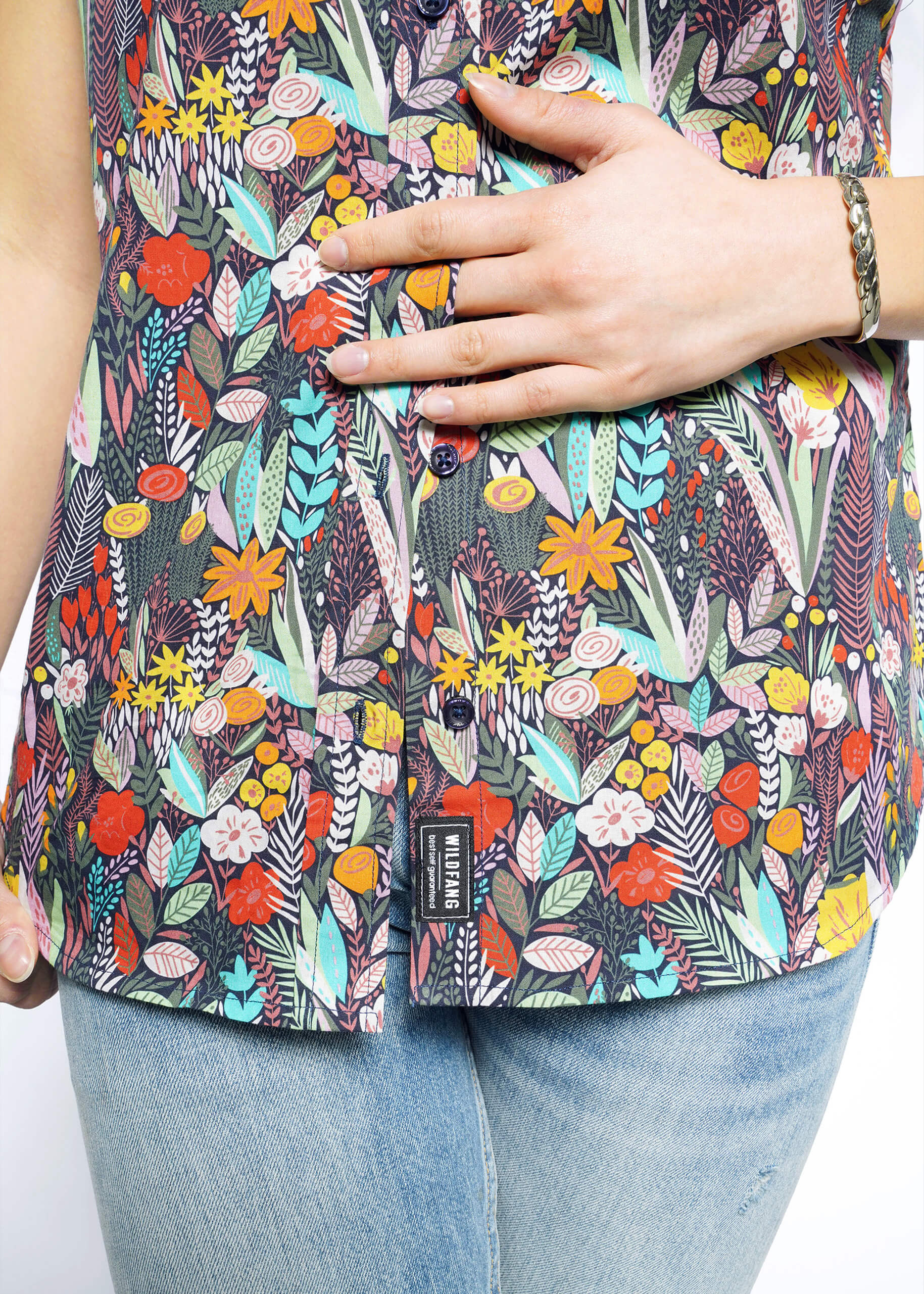 A person in Wildfangs The Essential Button Up, featuring a colorful floral pattern with red, yellow, orange, and blue blooms on breathable cotton. Paired with light blue jeans and a bracelet on the wrist, they rest a hand on their midsection.