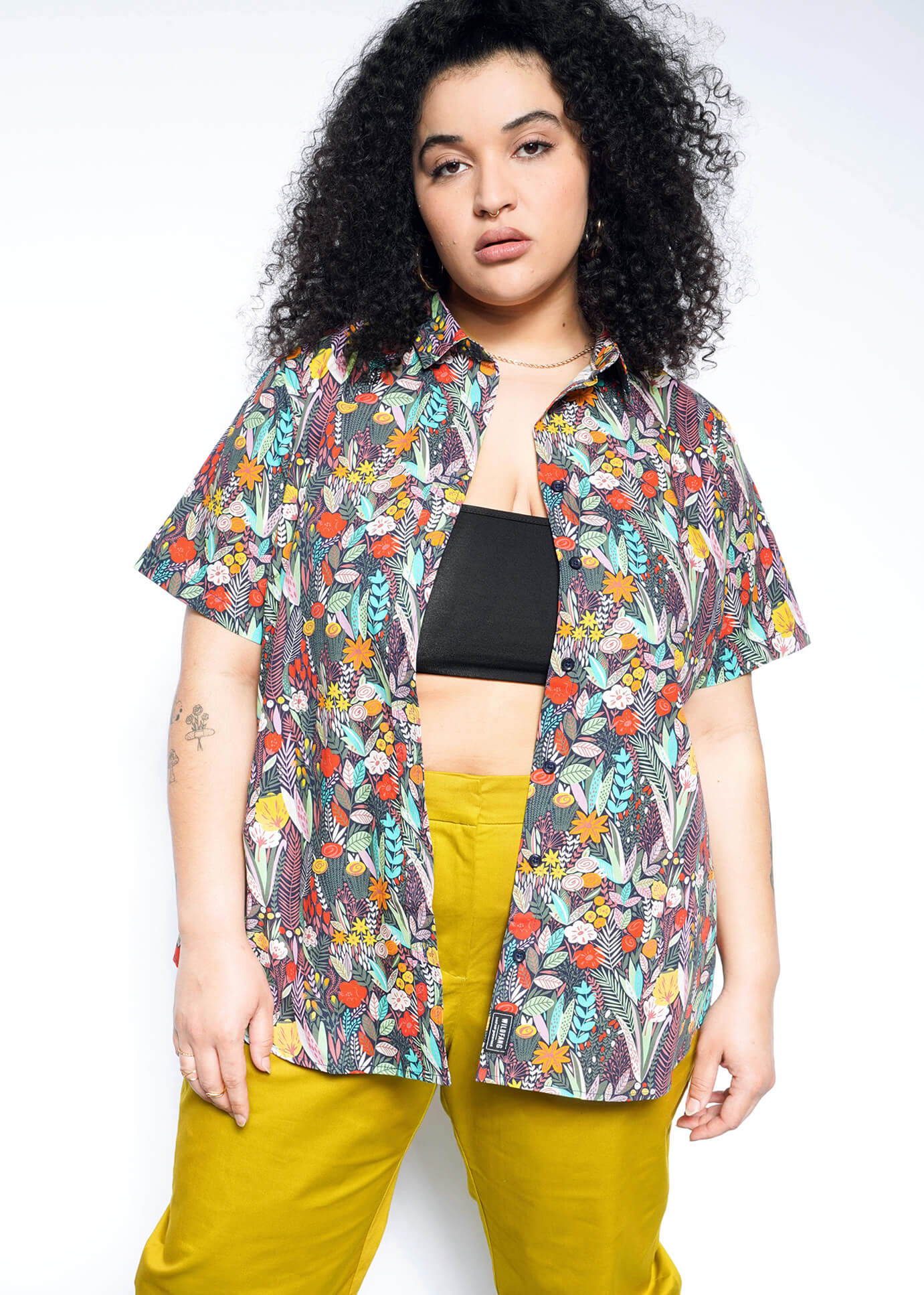 Wearing The Essential Button Up, a colorful floral shirt, over a black top and bright yellow pants, someone with curly hair stands confidently against a white background. The breathable cotton of the shirt ensures comfort as they look at the camera.