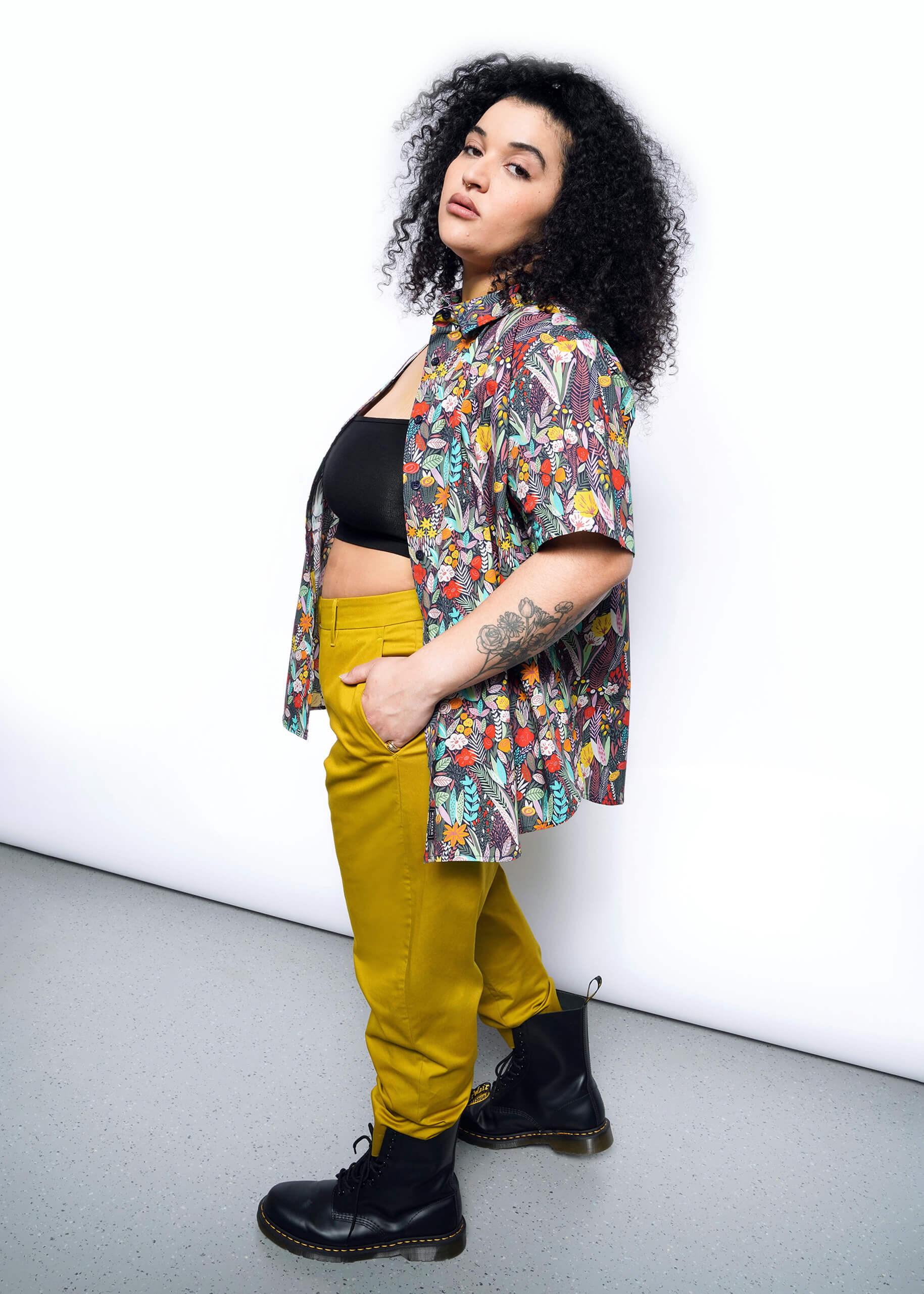 A person with curly hair confidently poses against a white background, donning the Wildfang floral-patterned The Essential Button Up and a black crop top. Their tailored yellow pants and black boots complete the look, while a tattoo on their arm adds an edgy touch.