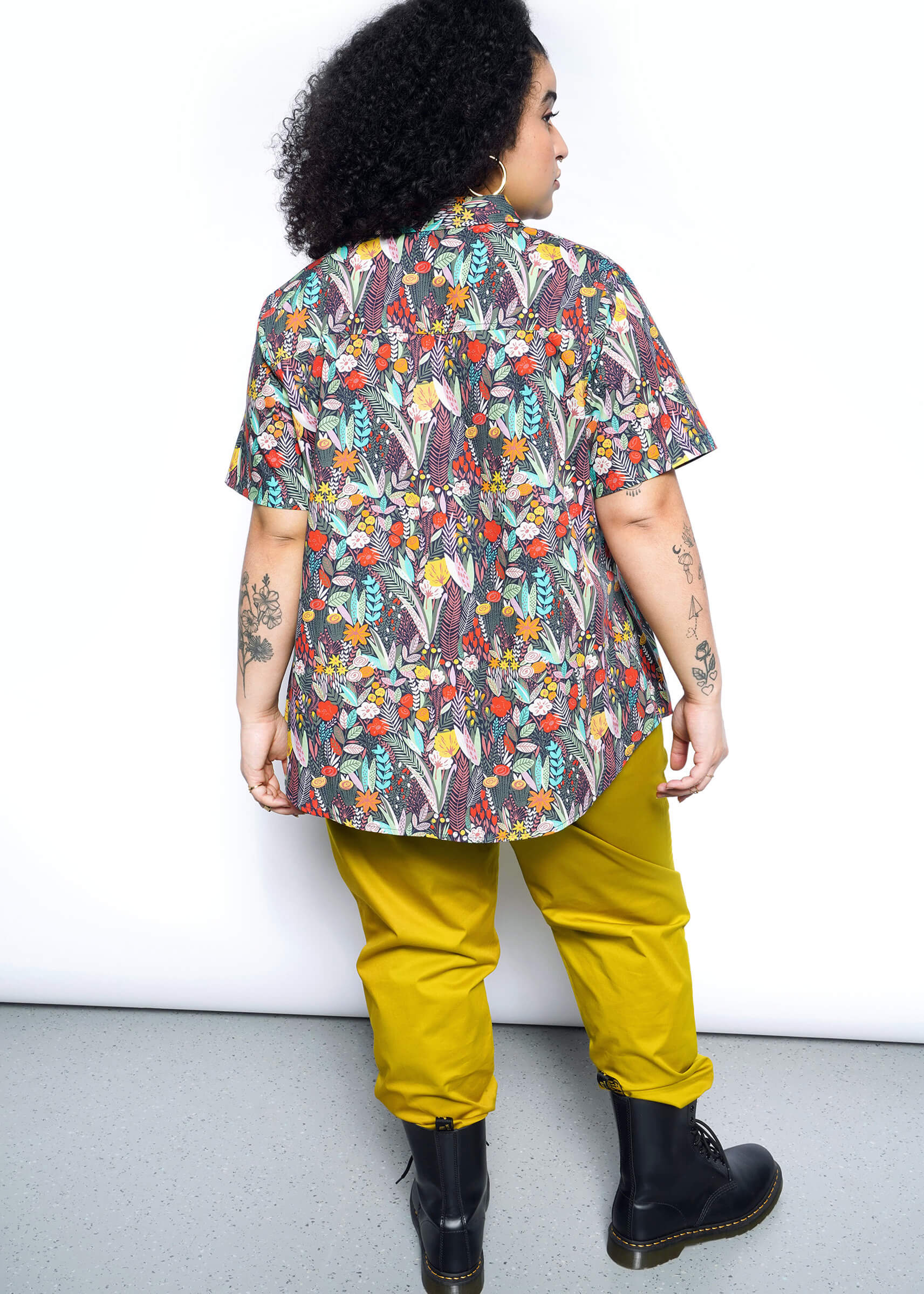 A curly-haired person, facing away, wears The Essential Button Up—a short-sleeve, floral-patterned shirt—in a tailored fit with mustard yellow pants. Standing in black boots on a light gray floor against a plain white wall, tattoos adorn their arms.