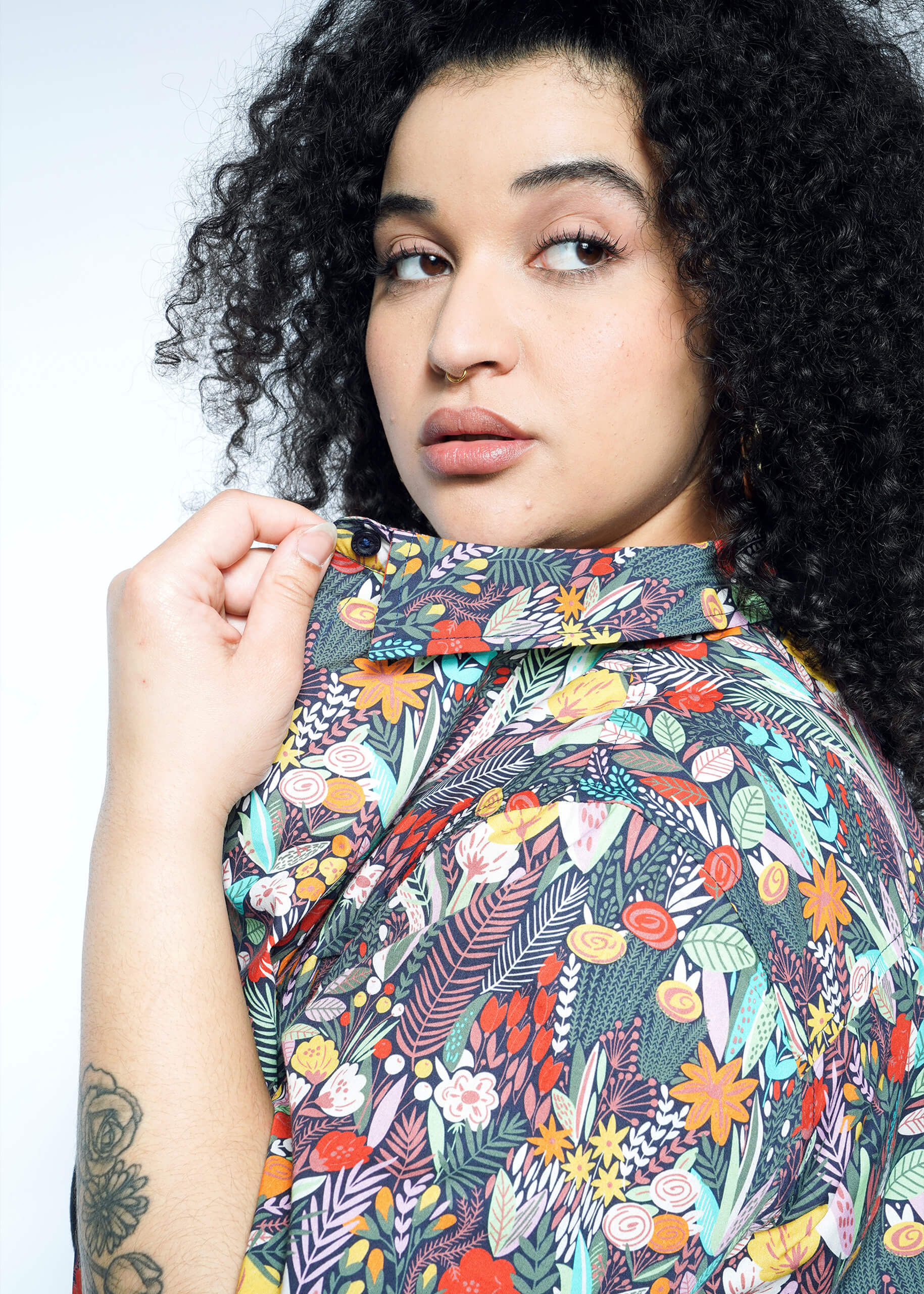 With curly hair, a person glances sideways in The Essential Button Up, a bold, floral-patterned shirt made of breathable cotton. They adjust the collar with one hand, revealing a flower tattoo on their forearm against the plain white background.