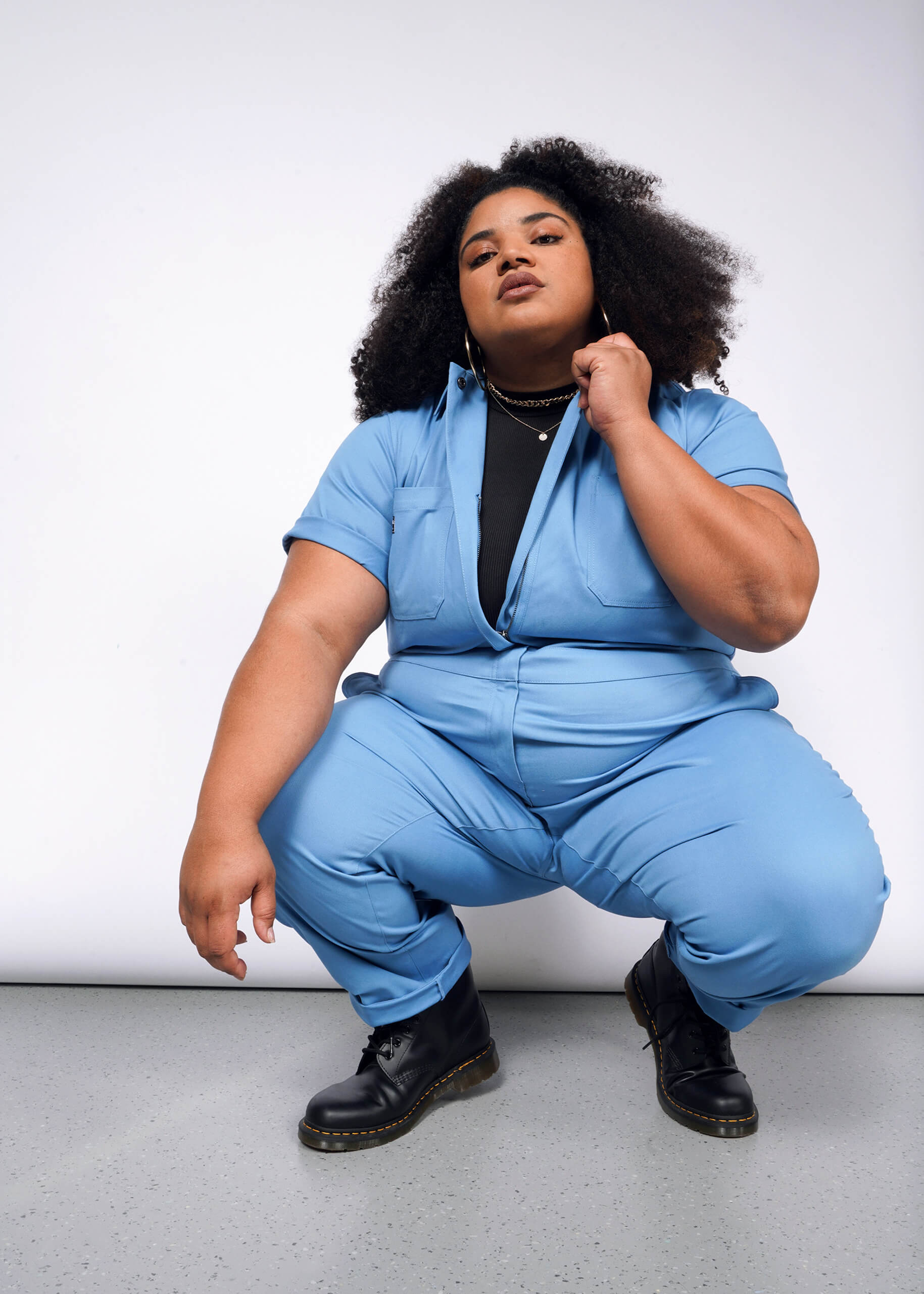 A person with curly hair confidently squats against a white background, wearing The Essential Coverall in light blue, which has a full-length front zipper and is made from stretch cotton. It features 9 pockets for style and functionality and is worn over a black top with black boots.