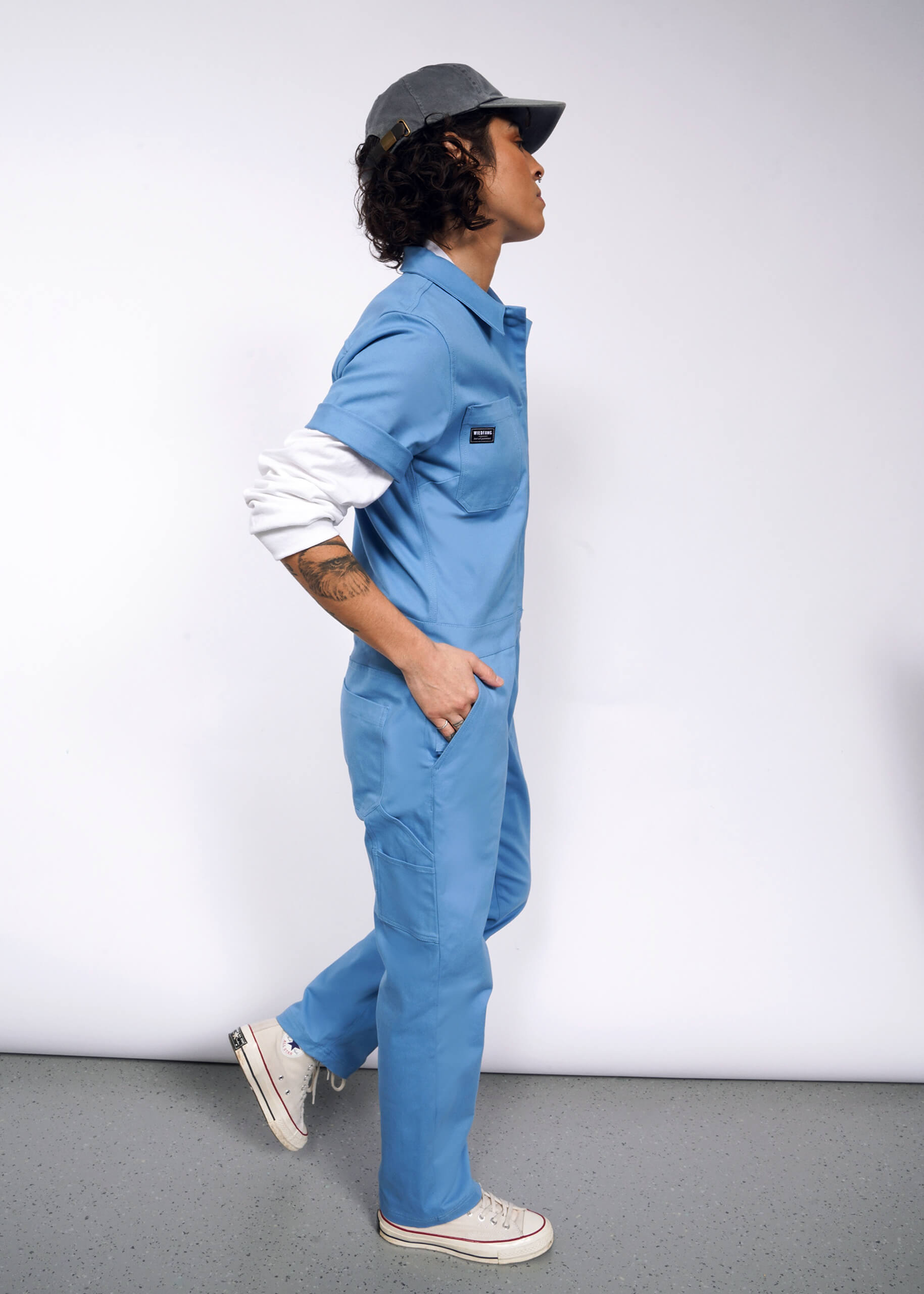 A person wearing The Essential Coverall stands in profile against a white backdrop. Featuring 9 pockets and a full-length front zipper, the stretch cotton blue jumpsuit pairs with a gray cap and white high-tops. One hand in their pocket, they glance sideways with flowing curls and a neutral expression.