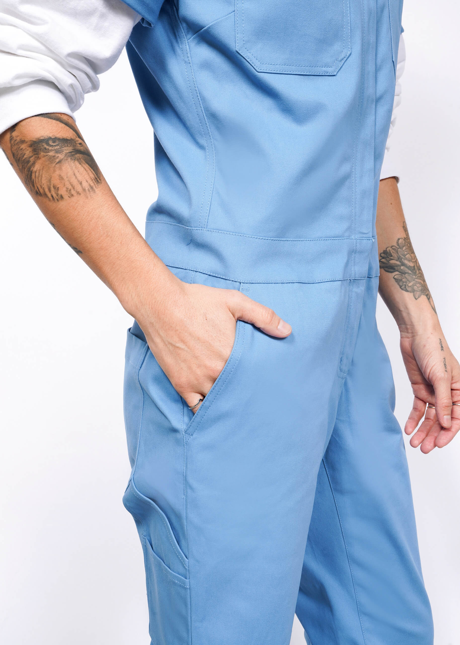 Dressed in The Essential Coverall, a person showcases a blue jumpsuit with a full-length front zipper and stretch cotton fabric. Their tattooed hand rests in their pocket against a plain white background.