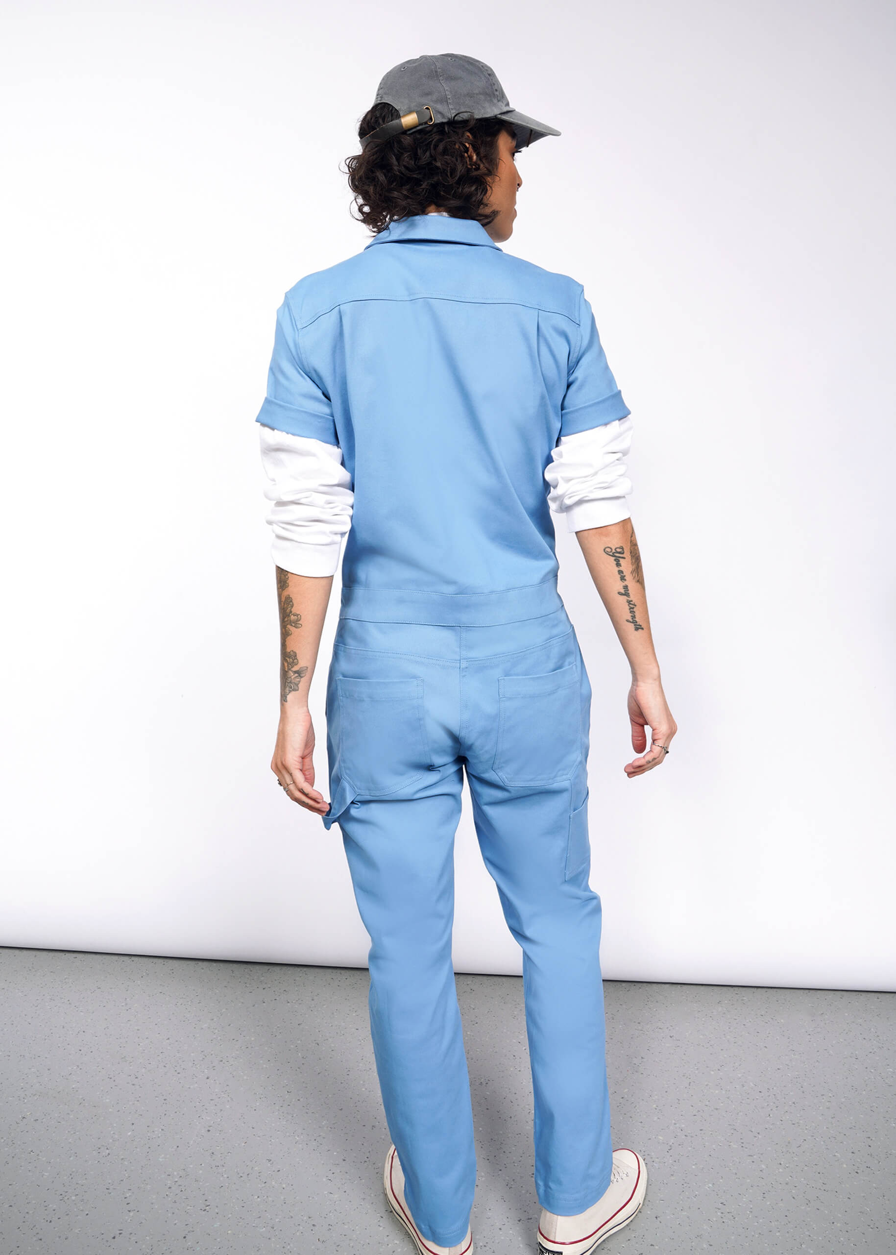 A person with curly hair, wearing The Essential Coverall in light blue with a full-length front zipper and a gray cap, faces away from the camera. Tattoos adorn their arms against a neutral backdrop. The stretch cotton of the coverall likely provides comfort and flexibility.