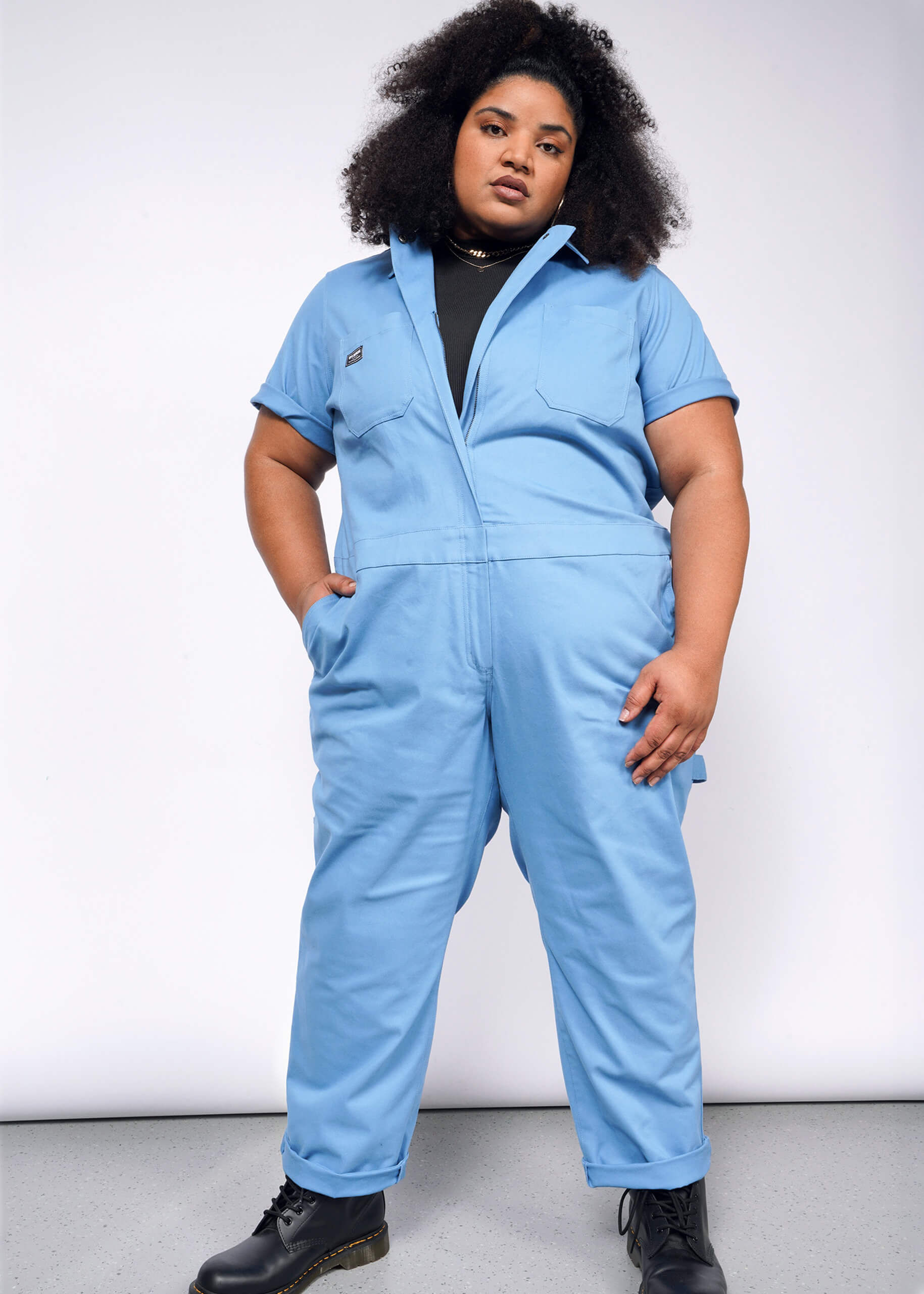 Against a plain backdrop, a person confidently wears The Essential Coverall, a light blue stretch cotton jumpsuit with a full-length front zipper. Paired with black boots and styled curly hair, the outfits 9 pockets offer practicality to their poised look.