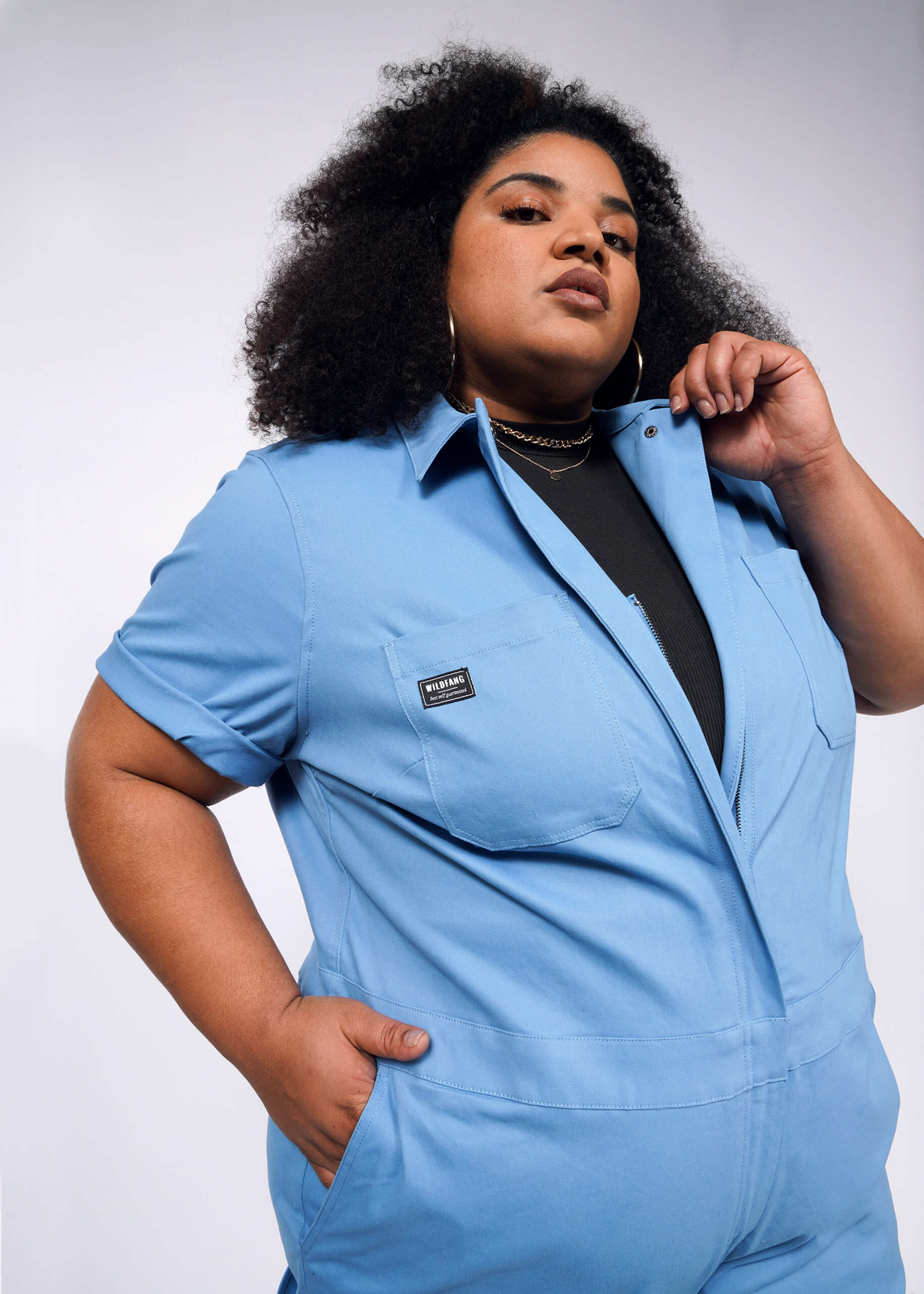 Dressed in The Essential Coverall, a stylish blue stretch cotton jumpsuit with a front zipper and nine convenient pockets, an individual poses confidently against a plain gray backdrop. Their curly hair frames their face as they casually hold the collar with one hand.