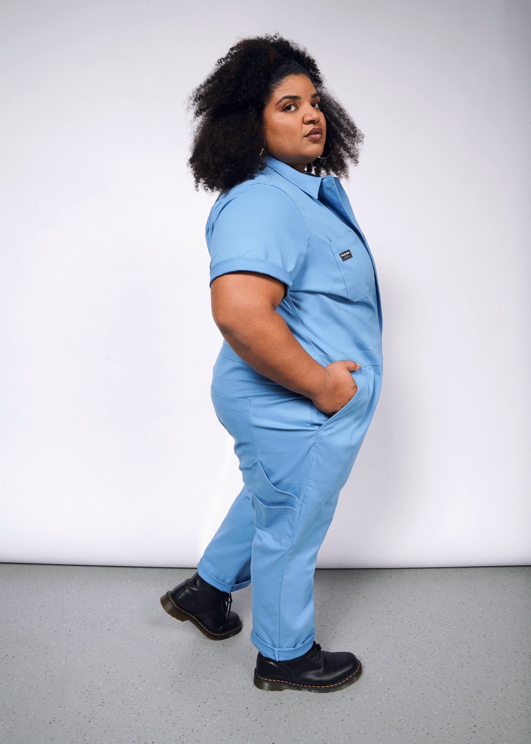 Against a plain white background, a person stands confidently in The Essential Coverall—light blue stretch cotton with a full-length front zipper and nine pockets. With black boots and curly hair adding flair, their hands are casually tucked in.