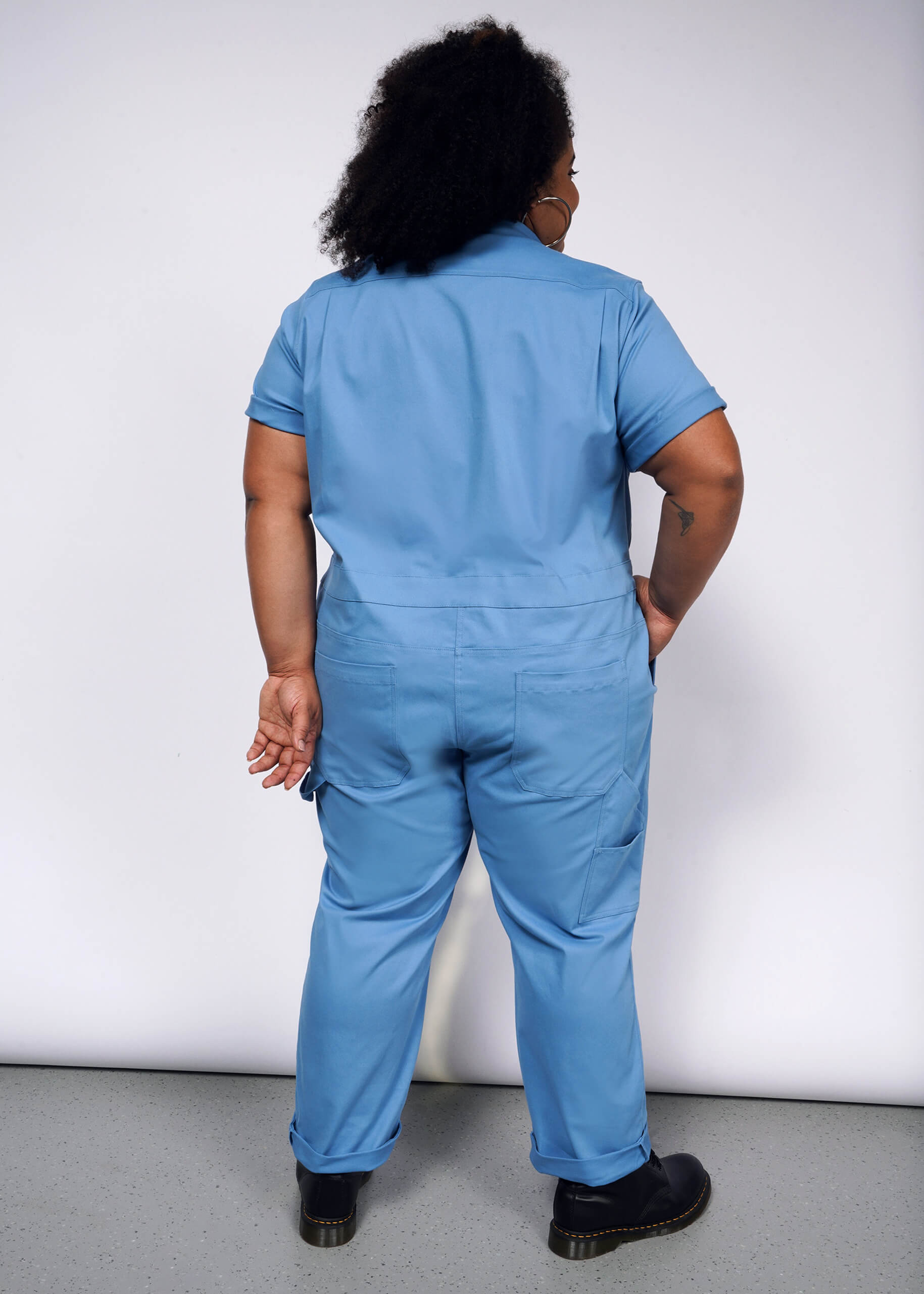 Facing away from the camera with short curly hair, a person wears The Essential Coverall in light blue stretch cotton featuring a full-length front zipper. Theyre paired with black shoes and hold an object in their right hand against a plain white backdrop.