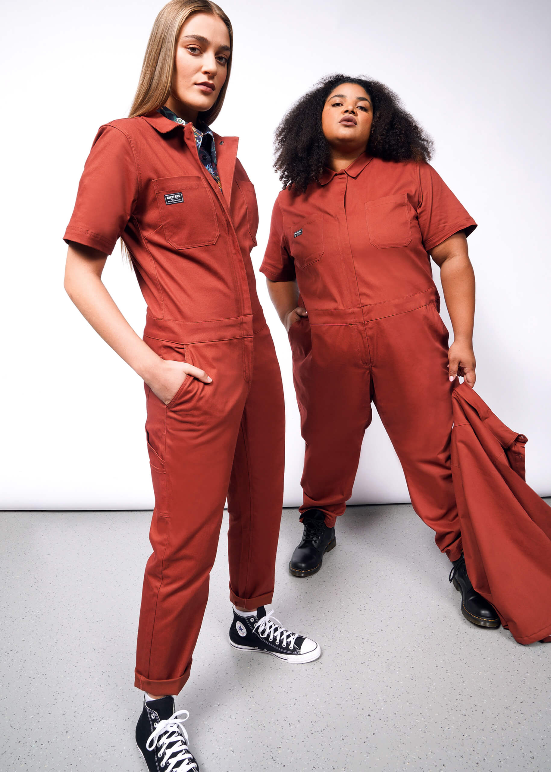 Two individuals confidently wear The Essential Coverall in rust, made from stretch cotton. One person has long hair, hands in pockets, and black sneakers; the other has curly hair, holds a jacket, and wears black boots. The relaxed fit with zipper front suits the simple background perfectly.