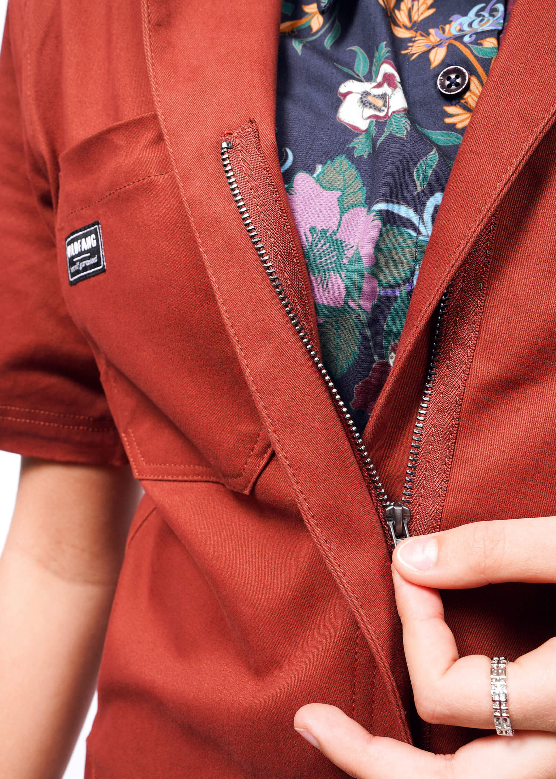 Dressed in The Essential Coverall with a rust hue and partially open zipper, revealing a floral patterned shirt underneath, the person enjoys the relaxed fit while holding the zipper. One hand is adorned with rings.