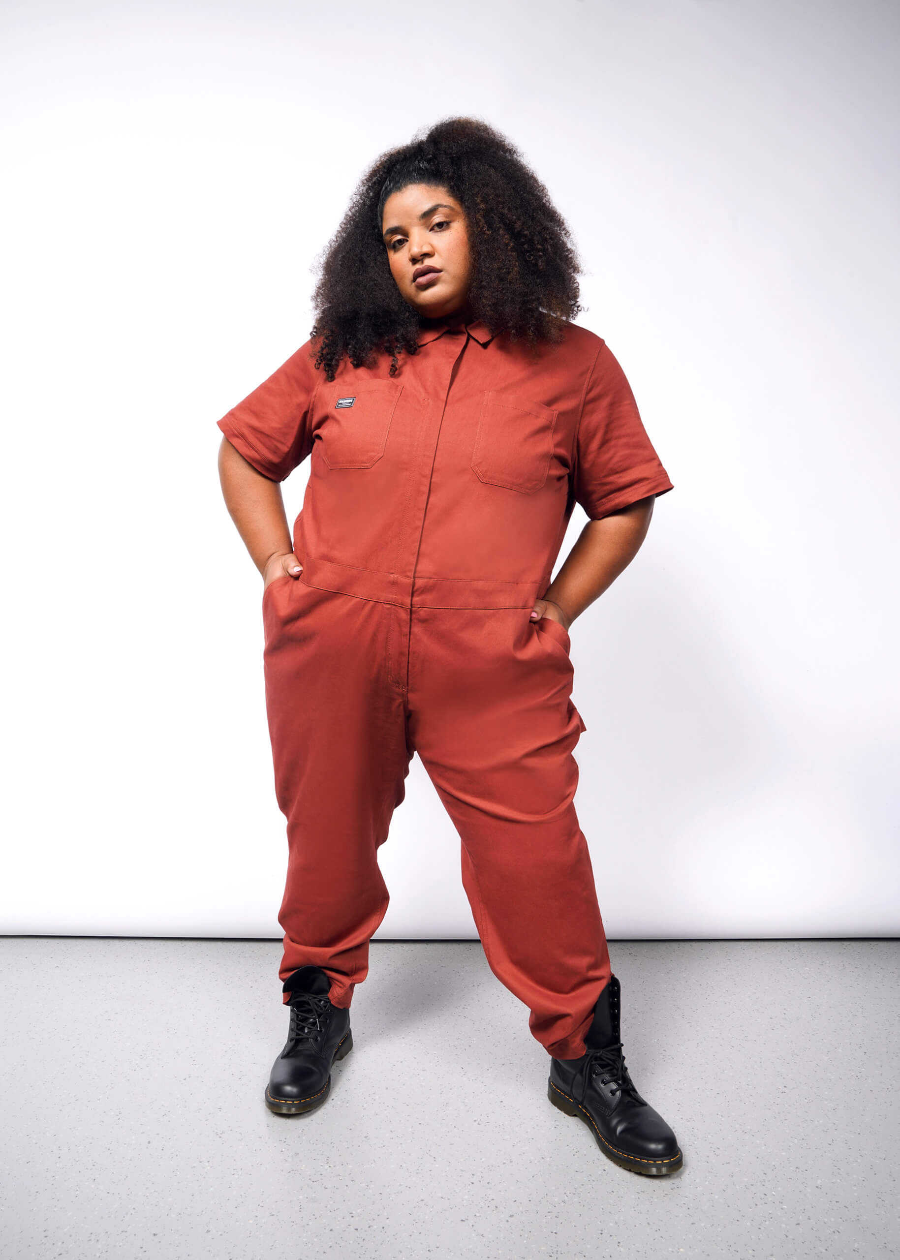 Wearing The Essential Coverall and black boots, a person with long curly hair stands confidently against a white background. The relaxed fit with stretch cotton hugs comfortably, and the zipper front adds style. With hands in pockets, they gaze steadily at the camera.