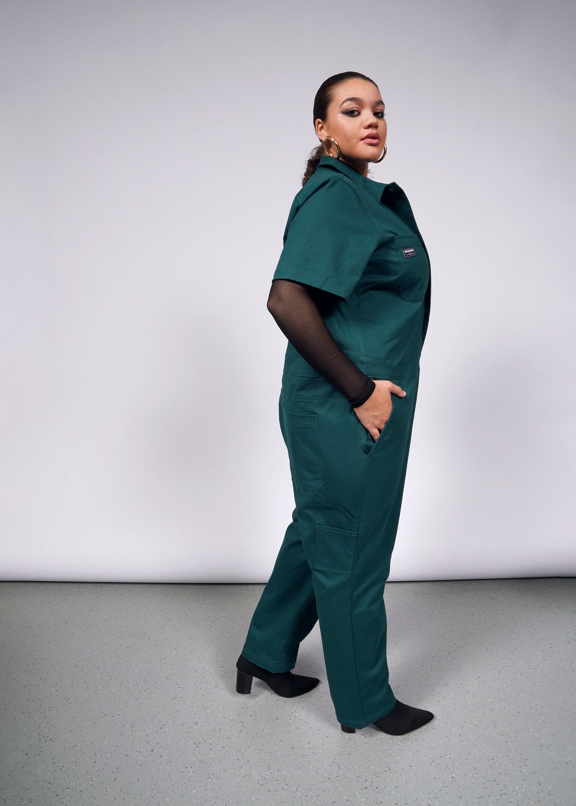 A person poses confidently in The Essential Coverall, a green jumpsuit crafted from soft brushed stretch cotton. It features short sleeves, side pockets, and a full-length front zipper. With one hand in a pocket and black boots on, the plain background enhances its color and design.