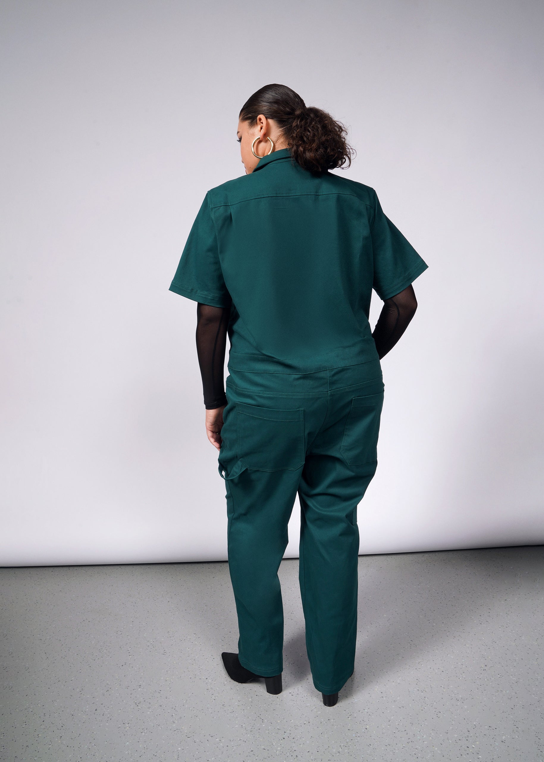 A person stands with their back to the camera wearing The Essential Coverall in dark green, featuring 9 pockets. The soft brushed stretch cotton fabric gently conforms to their form, while curly hair is tied back against a simple studio backdrop.