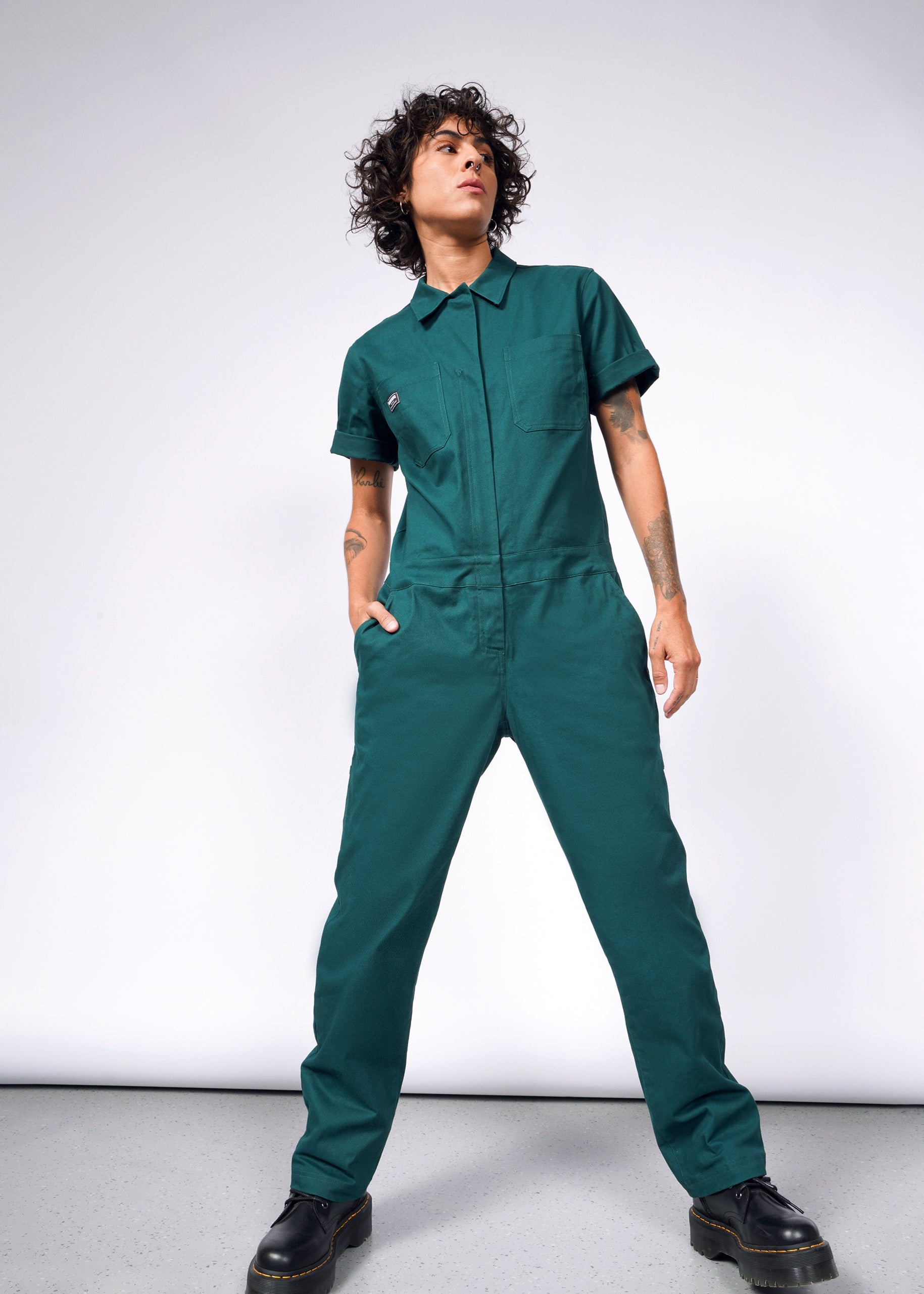 Wearing The Essential Coverall in soft brushed stretch cotton, a person stands confidently. It features short sleeves, rolled-up pant legs, a full-length front zipper, and an oversized fit. Their curly hair and black shoes complement the relaxed style against a plain white background.