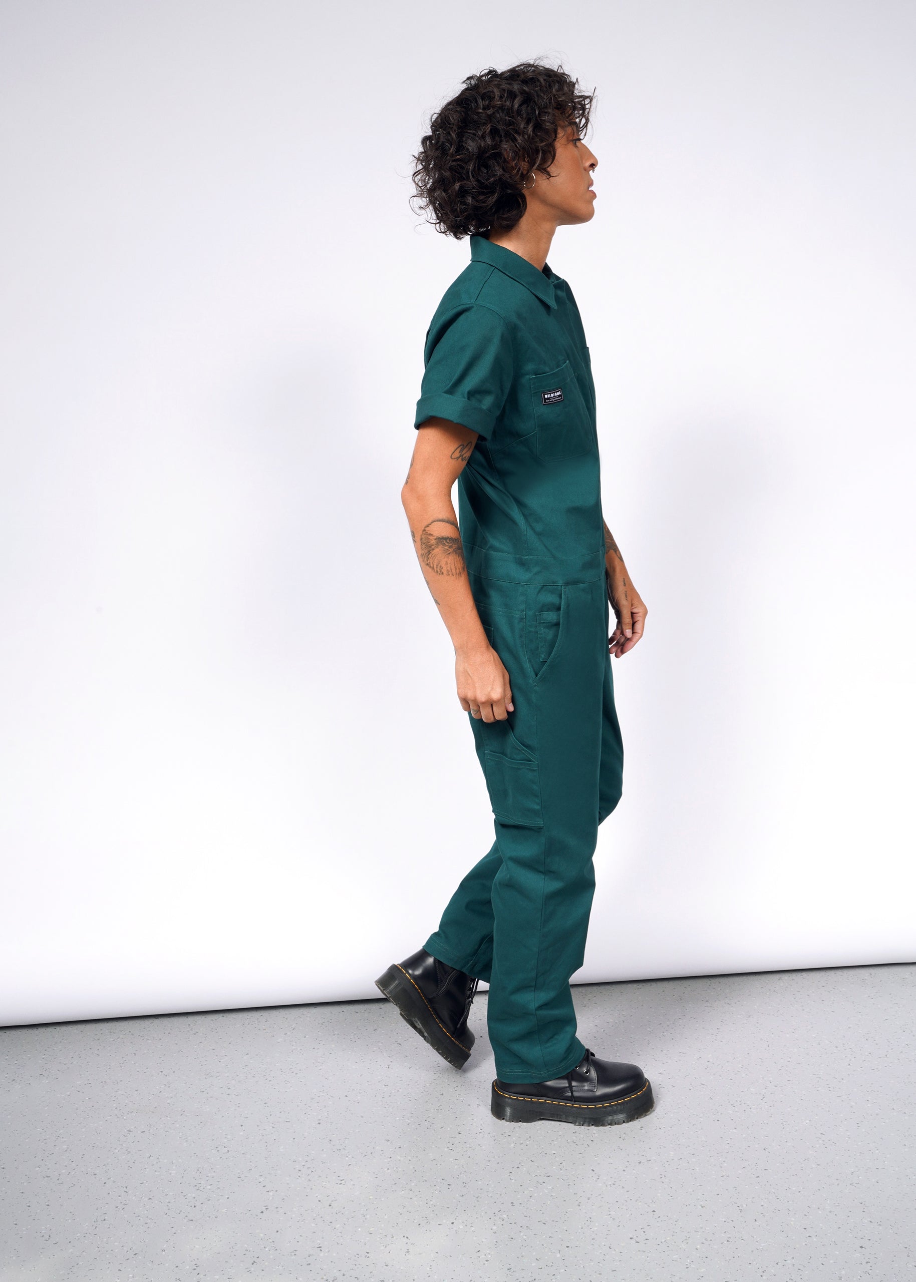 A person with curly hair stands sideways in The Essential Coverall, a green jumpsuit with a front zipper, paired with black boots. The soft brushed stretch cotton outfit highlights their left arm tattoos against a plain white background.