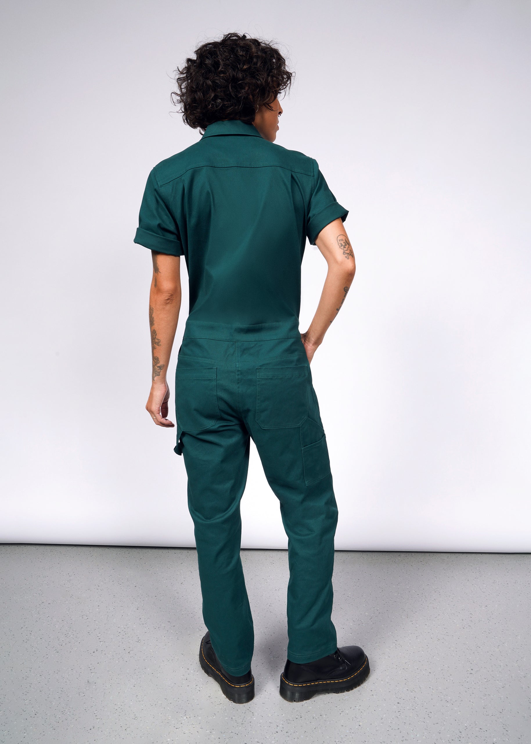 Against a light gray wall, a person with curly hair wears The Essential Coverall in green stretch cotton. The coverall boasts 9 pockets and a full-length zipper and is paired with black boots. Their tattooed arms are relaxed, giving an informal vibe.