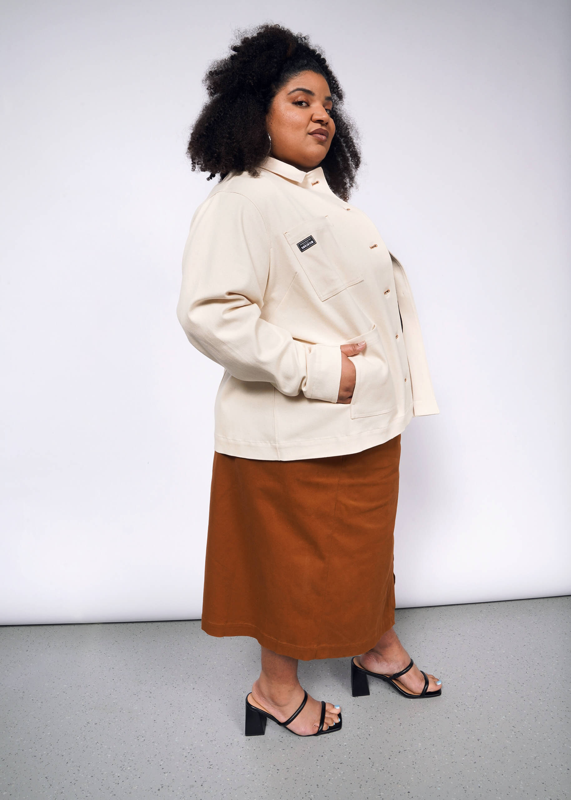 A person with curly hair confidently wears The Essential Denim Utility Jacket with deep pockets, paired with a brown skirt and black heeled sandals, standing in a studio against a neutral background.