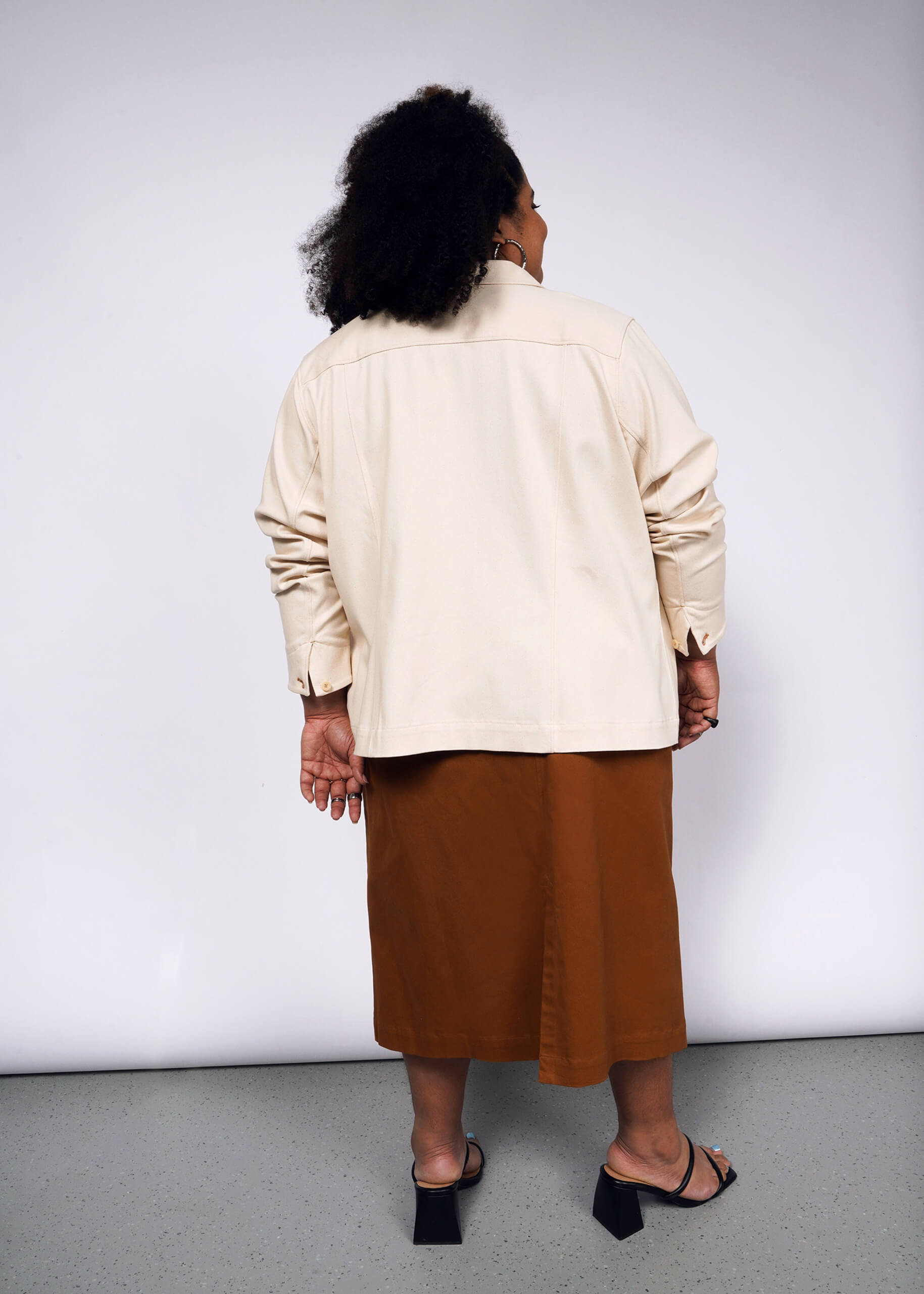 A person with curly hair stands with their back to the camera in a beige long-sleeve shirt, brown skirt, and black open-toe heels. Completing the look is The Essential Denim Utility Jacket, featuring deep pockets. The background is plain white.