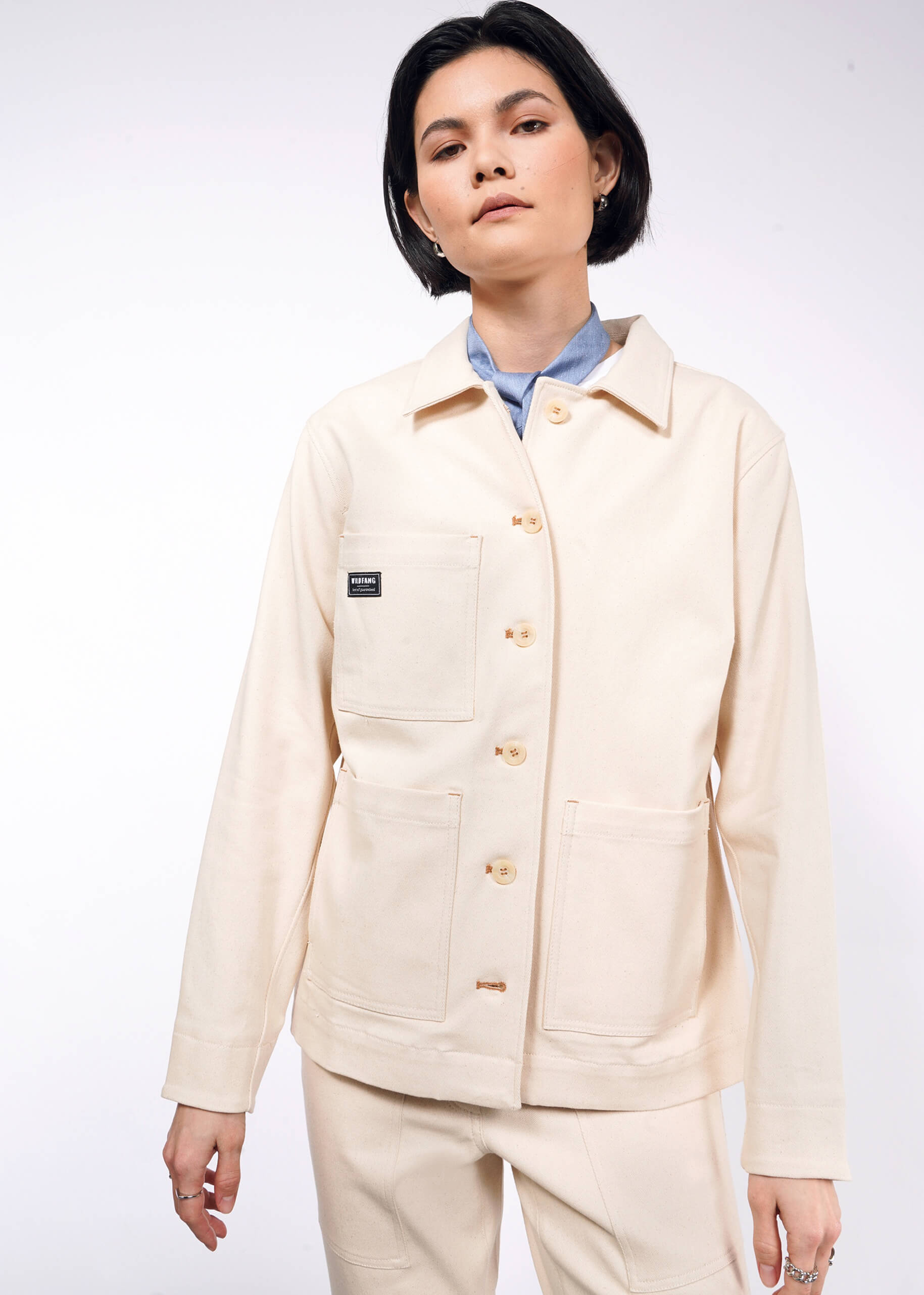 A person with short dark hair wears The Essential Denim Utility Jacket, known for its deep pockets and stretch comfort, over a light blue shirt. They stand against a plain white background, looking slightly to the side with a neutral expression.