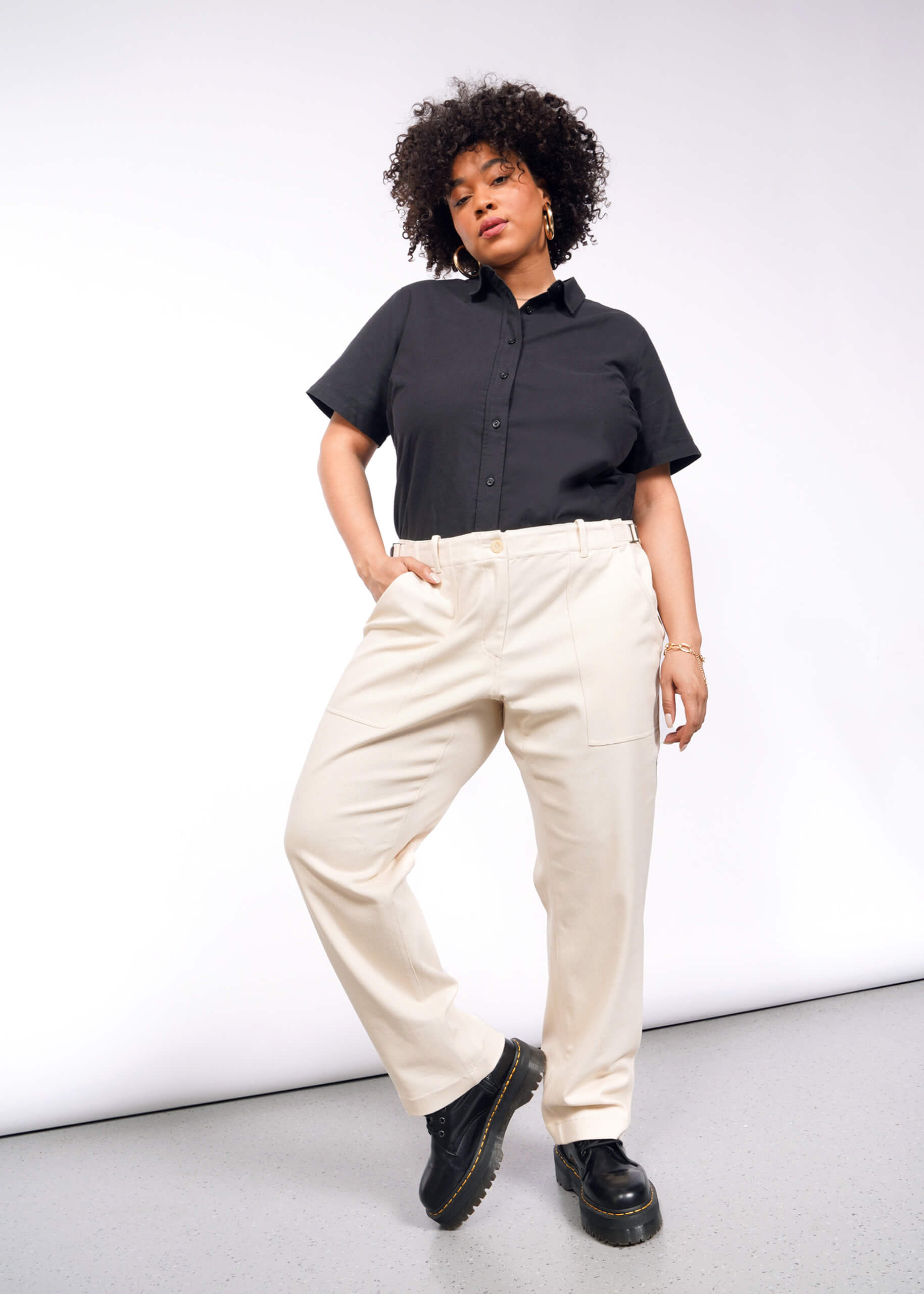 A person with curly hair stands confidently against a plain backdrop, sporting a stylish black short-sleeve button-up shirt and The Essential Denim Utility Pant in cream. The pants feature an adjustable waist and pair perfectly with their black platform shoes, as they look forward, hand in pocket.