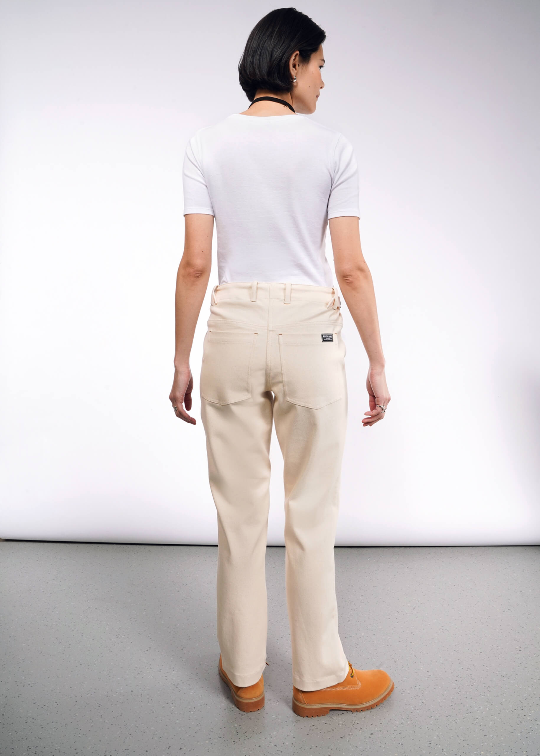 Rear view of a person with short dark hair wearing a white t-shirt, The Essential Denim Utility Pant in beige with an adjustable waist, and brown shoes, standing against a plain light-colored background.