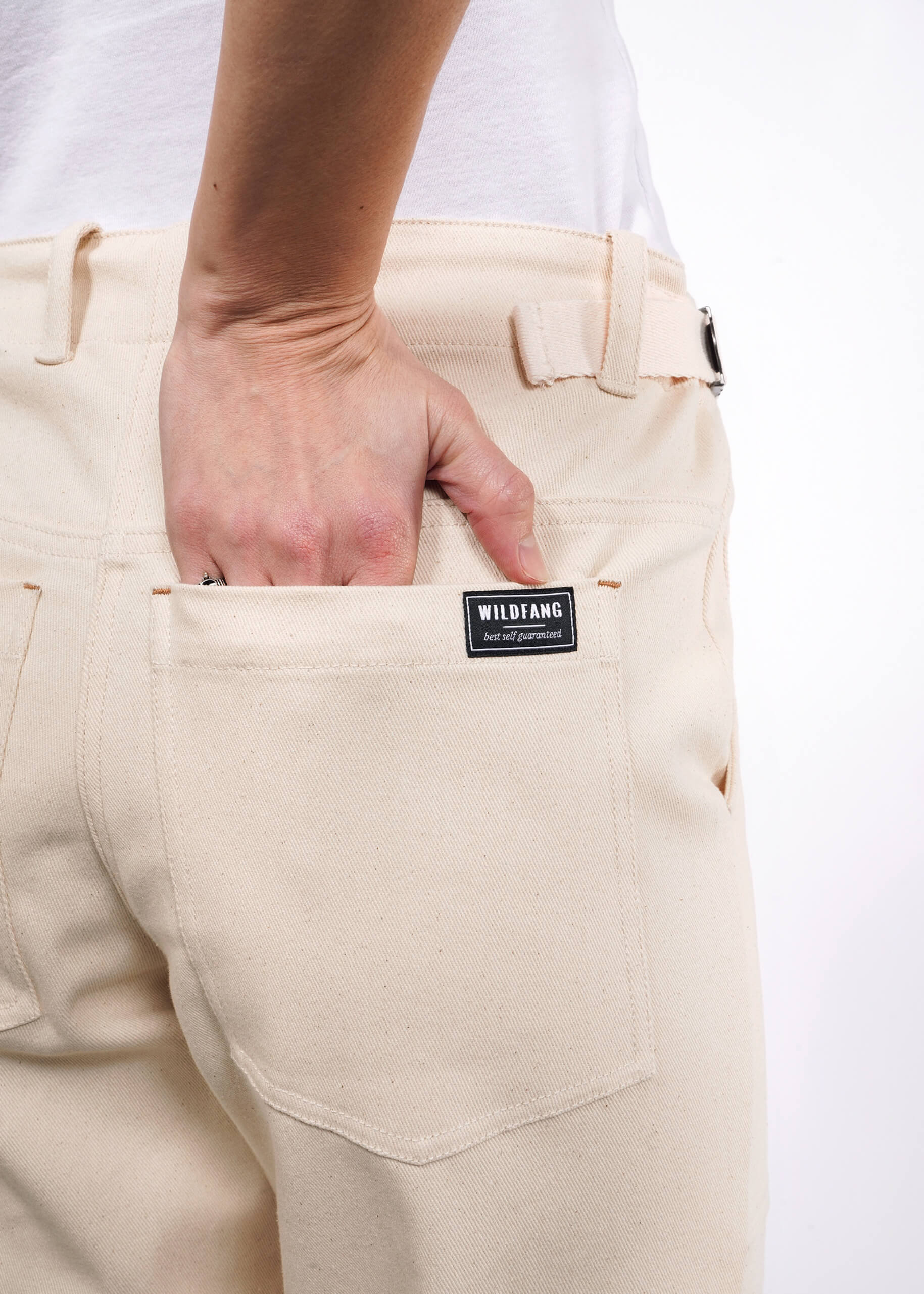 Close-up of someone in cream-colored The Essential Denim Utility Pant featuring a black WILDFANG label and stretch denim for added comfort as they slide their hand into the back pocket. Theyre styled with a white top against a simple white backdrop.