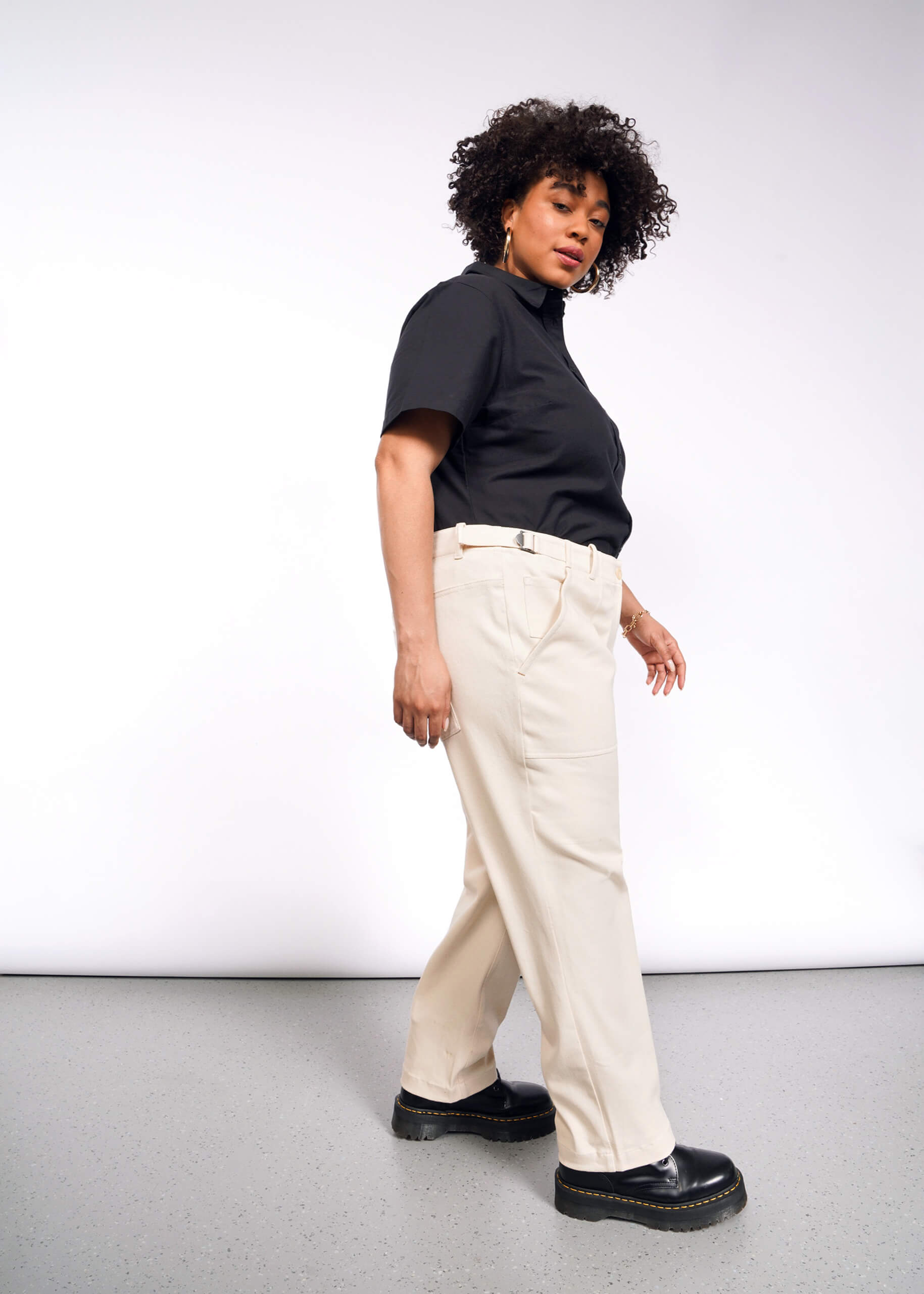 A person with curly hair stands sideways, wearing a black t-shirt and The Essential Denim Utility Pant in beige with an adjustable waist. Black boots complete the look against a plain white backdrop and gray floor in an indoor setting.