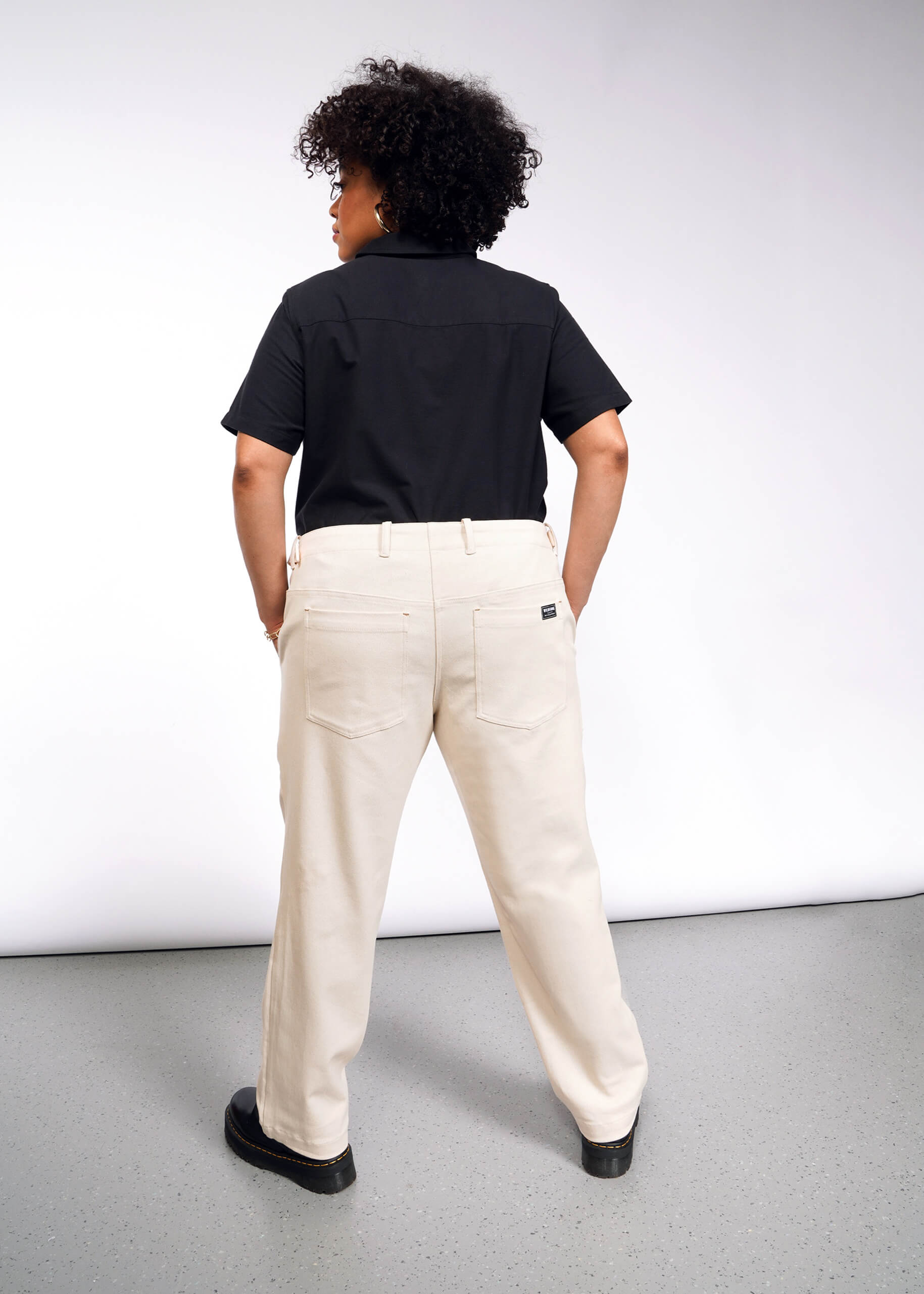 A person with curly hair stands facing away from the camera, wearing a black shirt, The Essential Denim Utility Pant in light beige with an adjustable waist, and black shoes. The background is a simple white and gray studio setting.