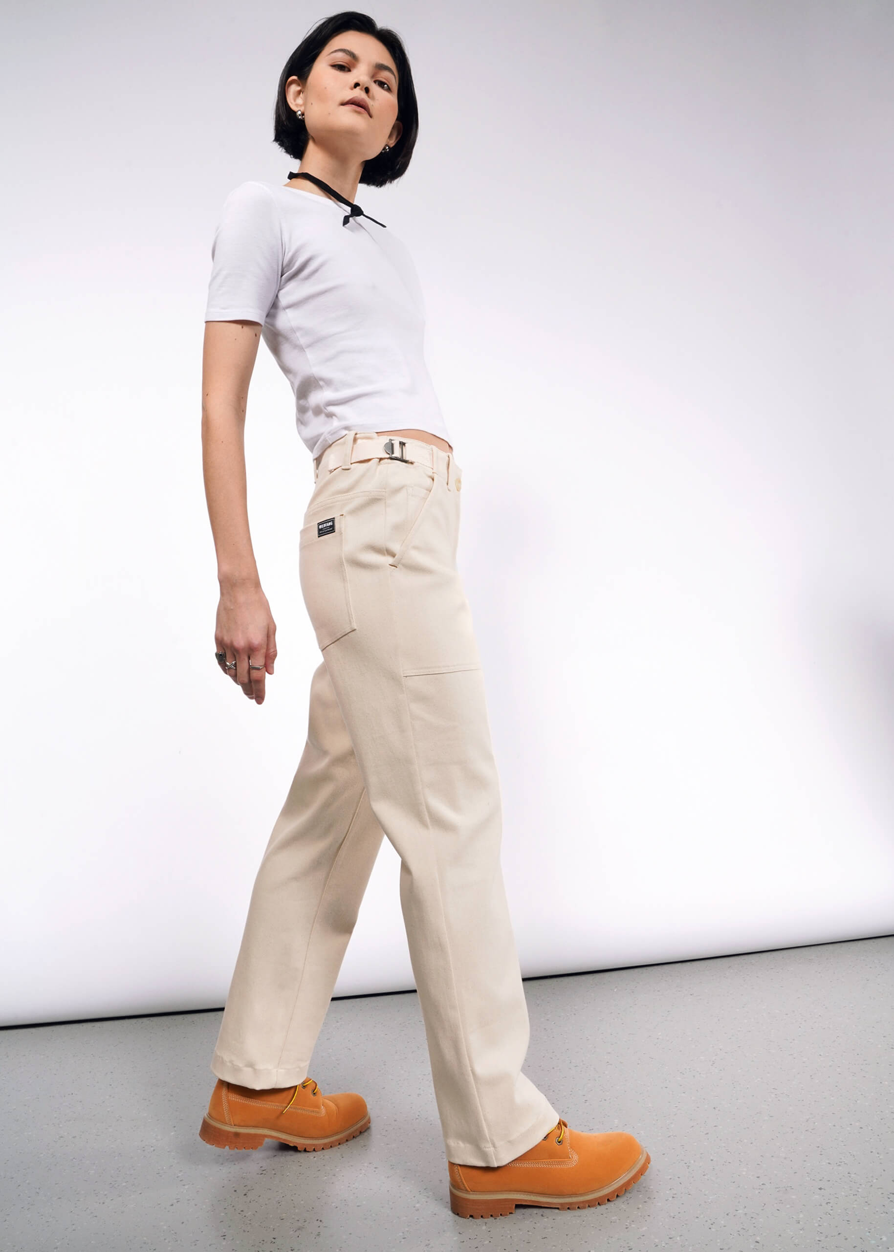 Someone in a white t-shirt, The Essential Denim Utility Pant, and tan shoes confidently poses against a plain white background. Their short, dark hair complements their casual yet stylish appearance, enhanced by the adjustable waist of the pants.