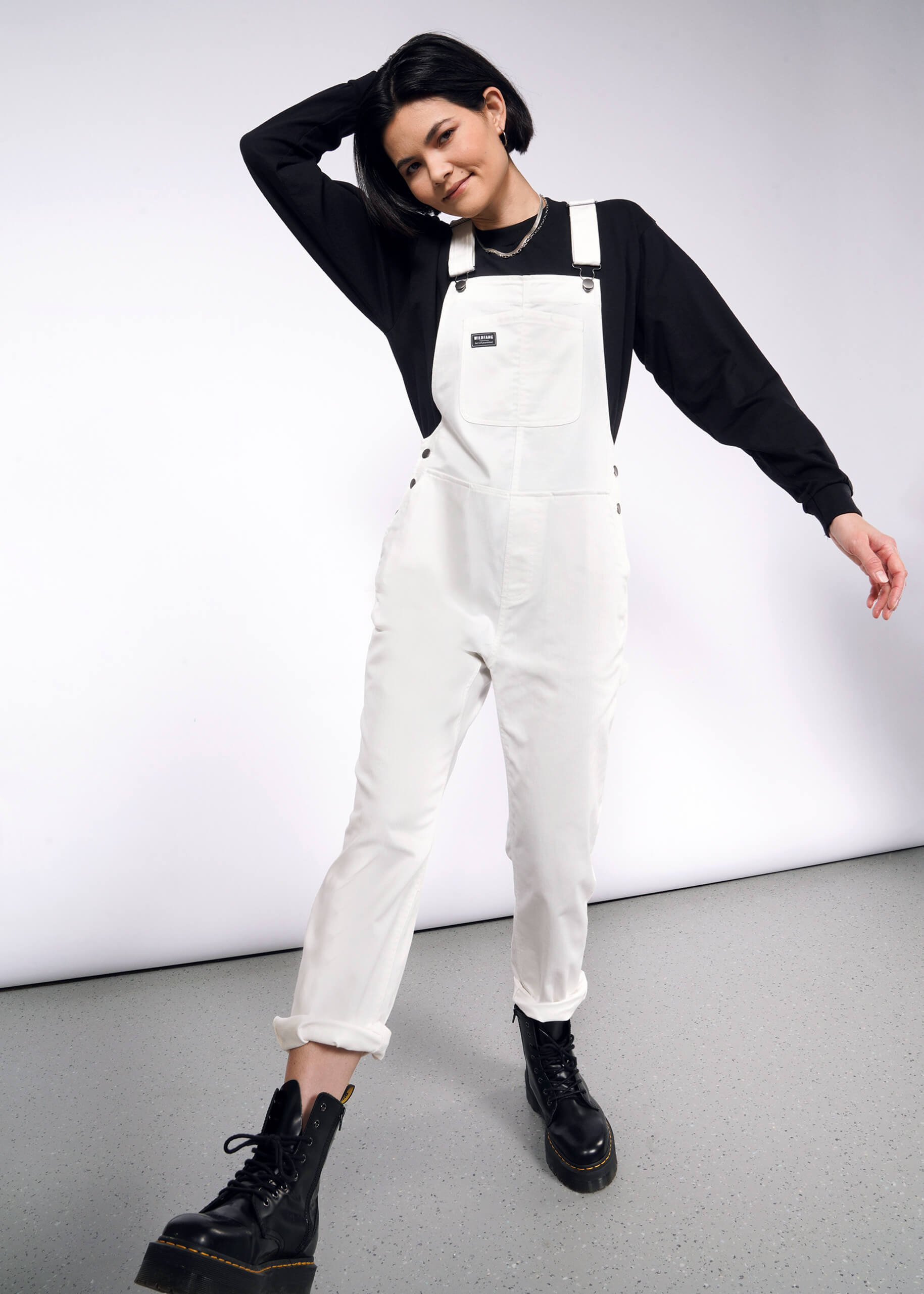 A person in The Essential Herringbone Overall with adjustable straps, a black long-sleeve shirt, and black boots poses playfully, one hand on their head, the other relaxed. They stand on a speckled gray floor against a white background.