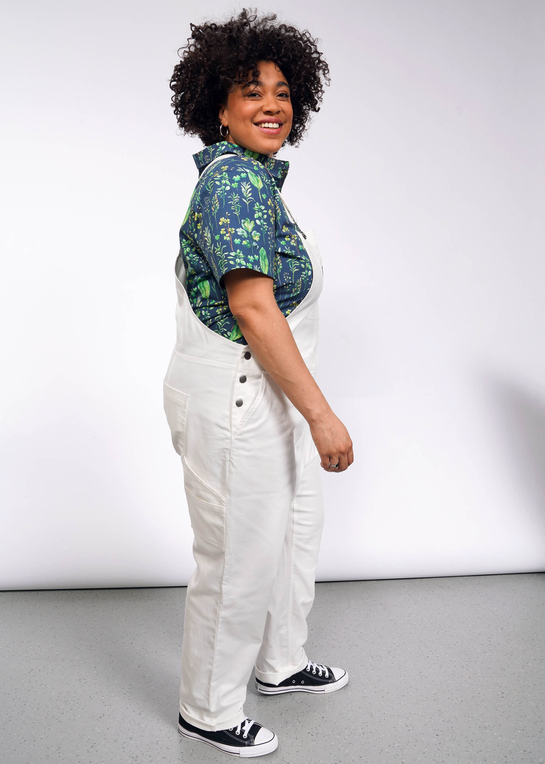 In a plain white studio, a curly-haired person smiles while standing sideways, wearing The Essential Herringbone Overall with deep pockets and adjustable straps over a blue patterned shirt, paired with black and white sneakers.