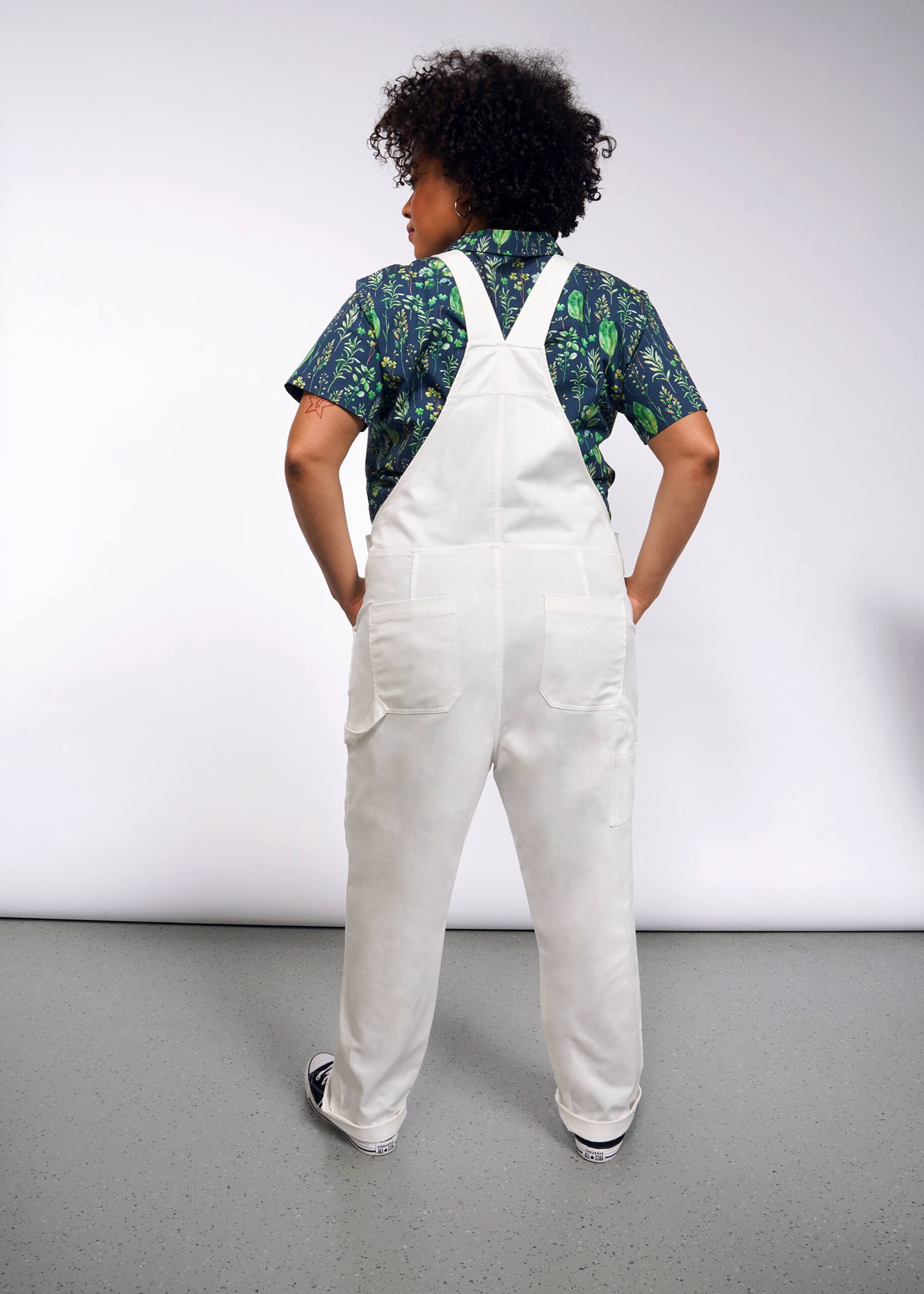 A person with curly hair facing away wears The Essential Herringbone Overall over a blue and green patterned shirt. On a gray speckled floor against a gray wall, their hands are tucked into the overalls deep pockets.