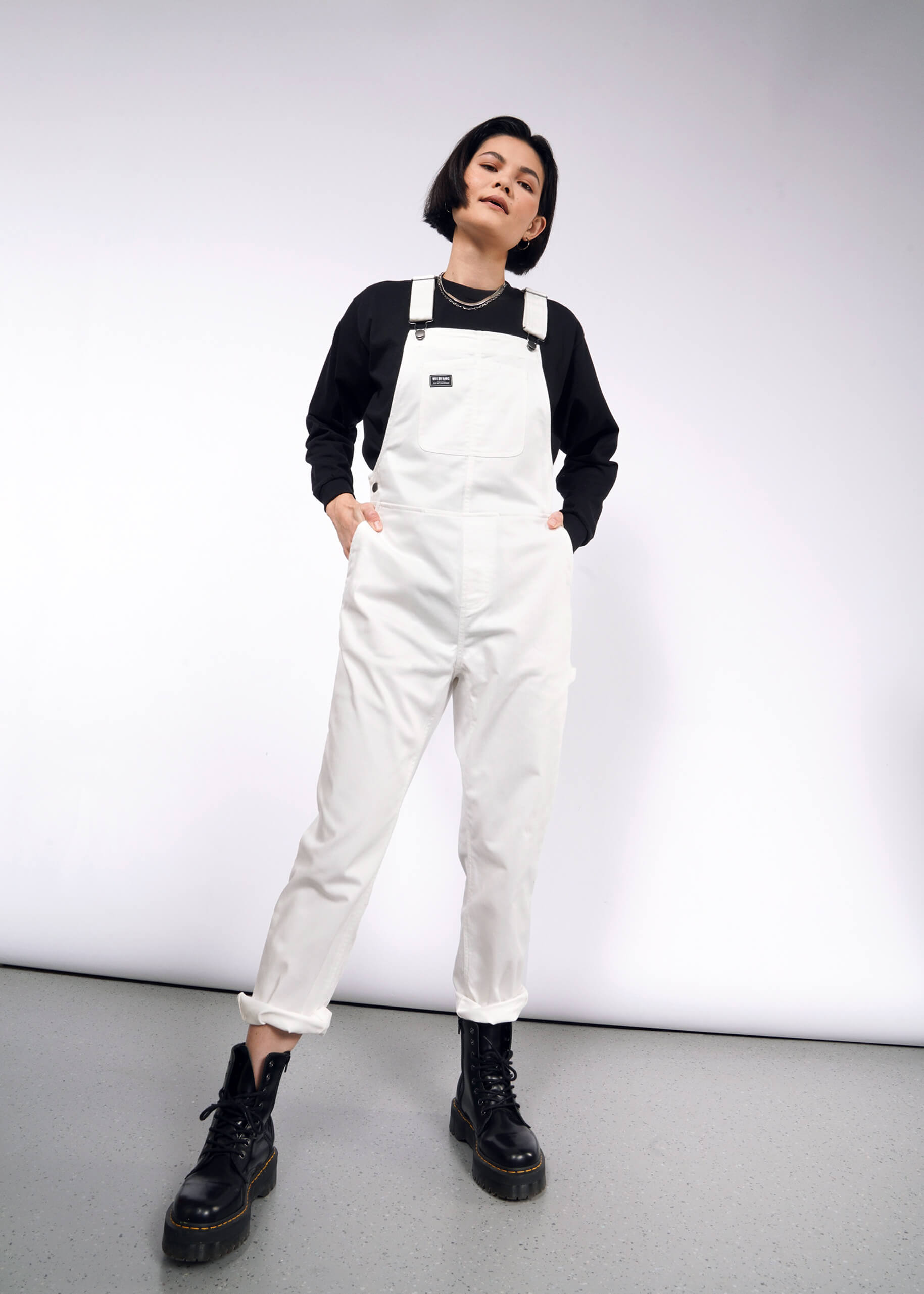 A person with short dark hair stands confidently against a plain background, wearing The Essential Herringbone Overall in white with adjustable straps and deep pockets, layered over a black long-sleeve shirt, and paired with black boots.