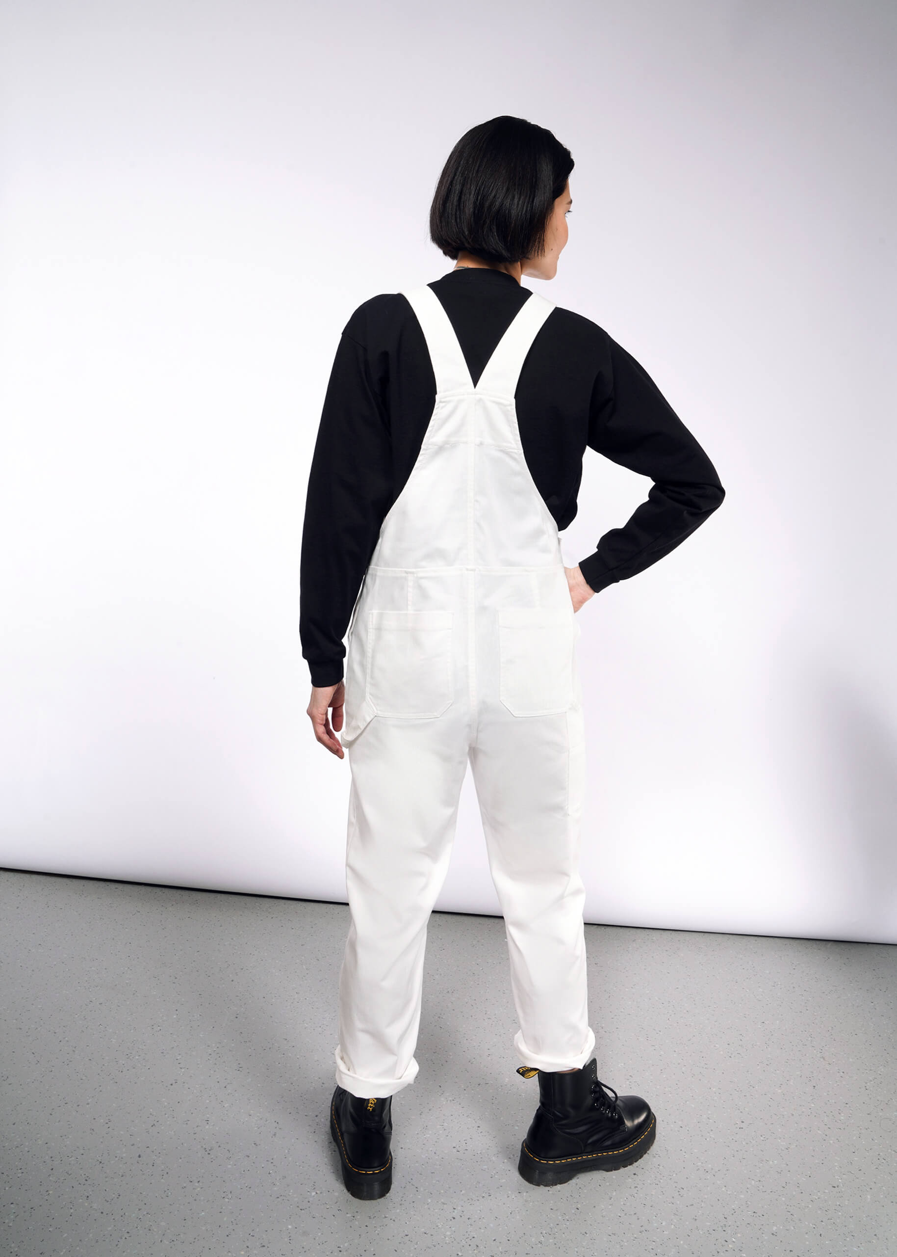 A person with short black hair in The Essential Herringbone Overall, adjustable straps, and a black long-sleeve shirt stands hand on hip in a white room with a gray floor, wearing black boots. They face away from the camera.