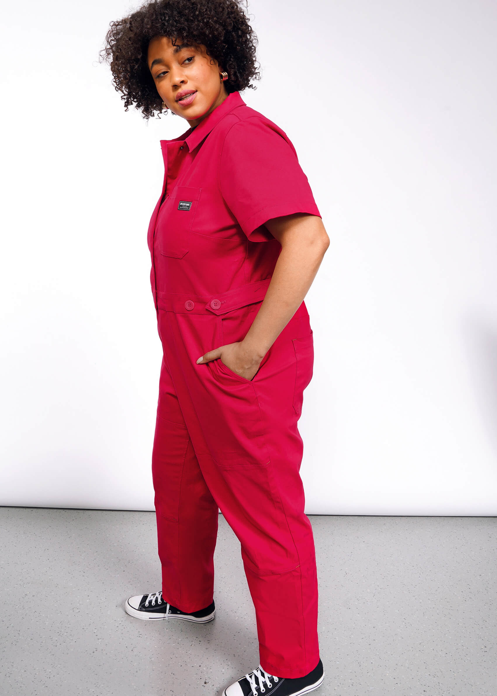 A person with curly hair wearing The Essential High Waisted Coverall in bright pink with deep pockets and black sneakers stands against a plain white background, hands in pockets, gazing confidently to the side.