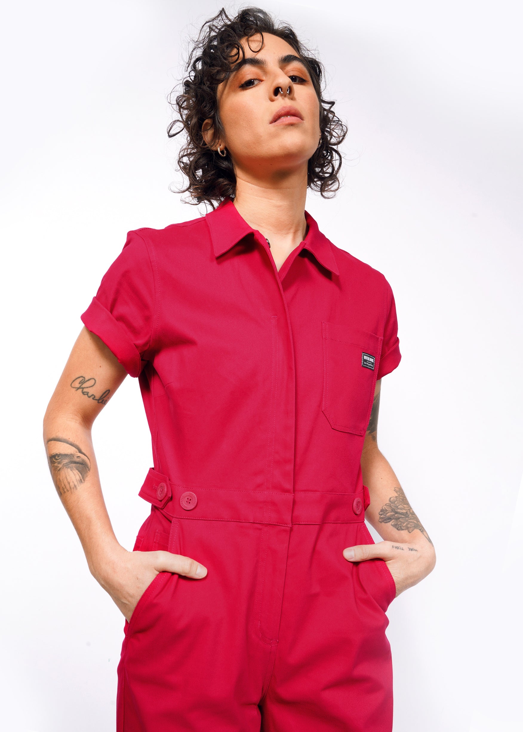 A curly-haired person poses in The Essential High Waisted Coverall in red, with hands in deep pockets. Tattoos are visible on both arms against a plain white background, highlighting the styles high-waisted fit.