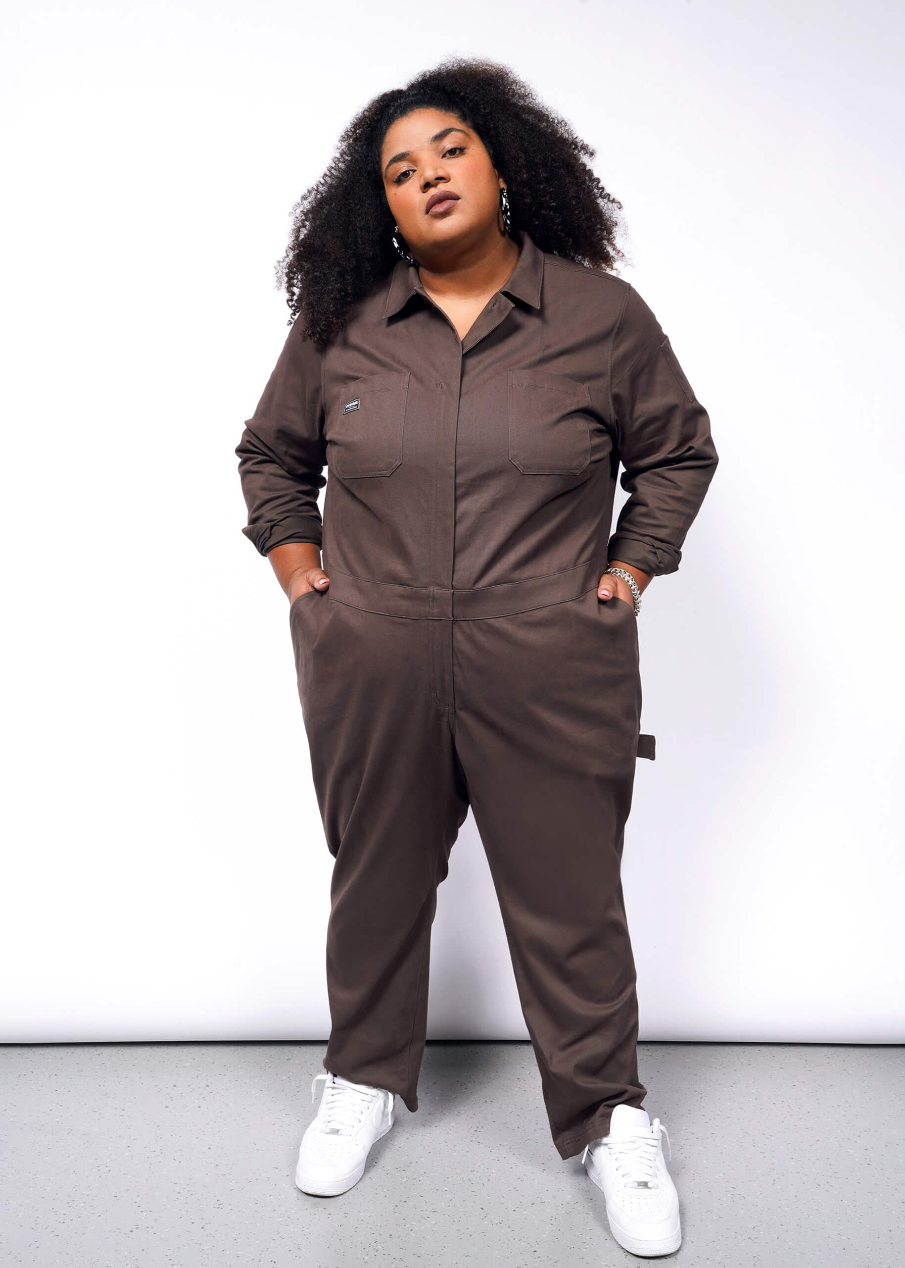 Someone with curly hair stands confidently in an oversized fit of The Essential Long Sleeve Coverall, made from brushed stretch cotton in brown, paired with white sneakers against a plain background. Their hands rest in the pockets of the jumpsuit.