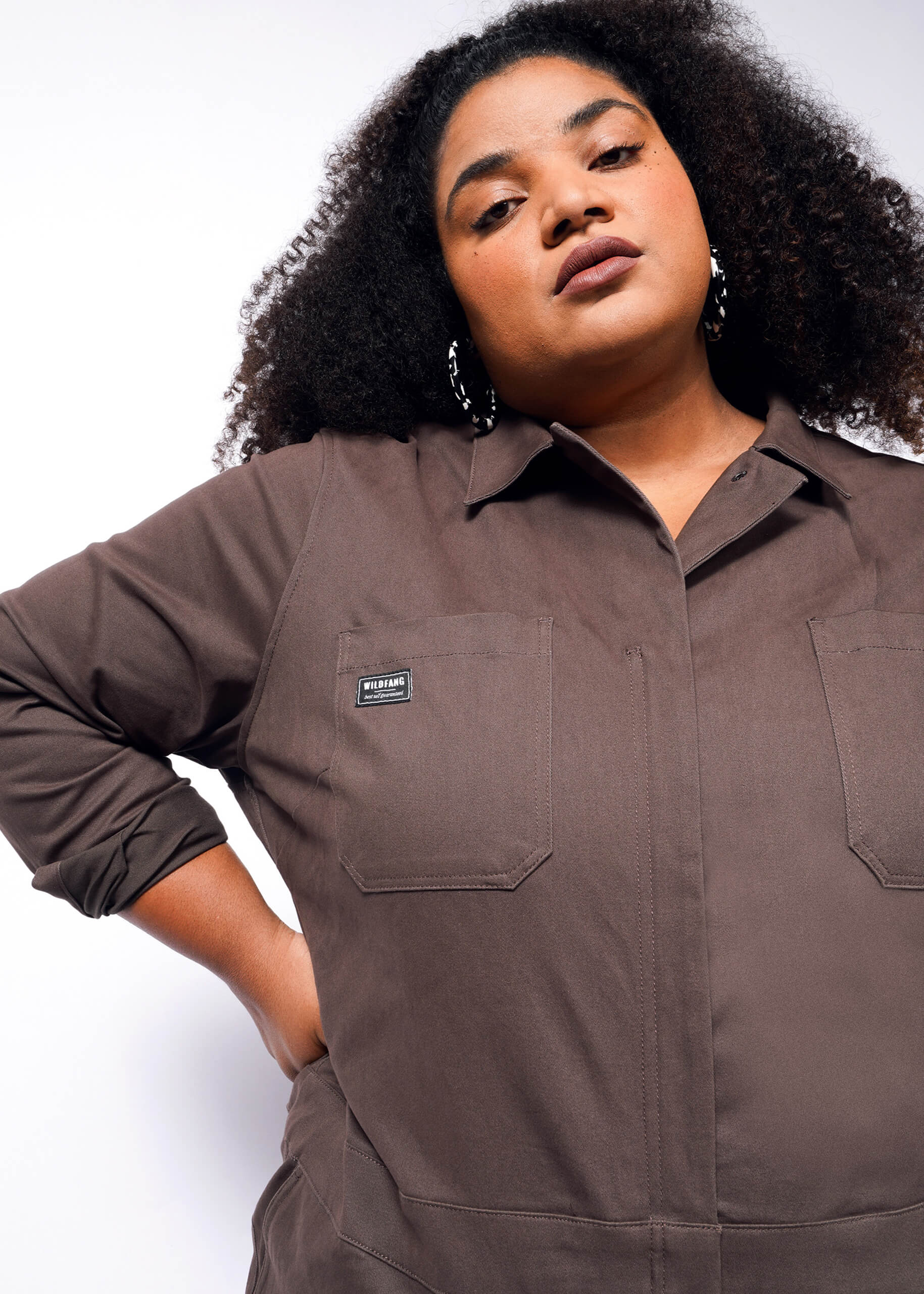 A person with curly hair confidently poses against a white background wearing The Essential Long Sleeve Coverall in brown, featuring brushed stretch cotton and two chest pockets.