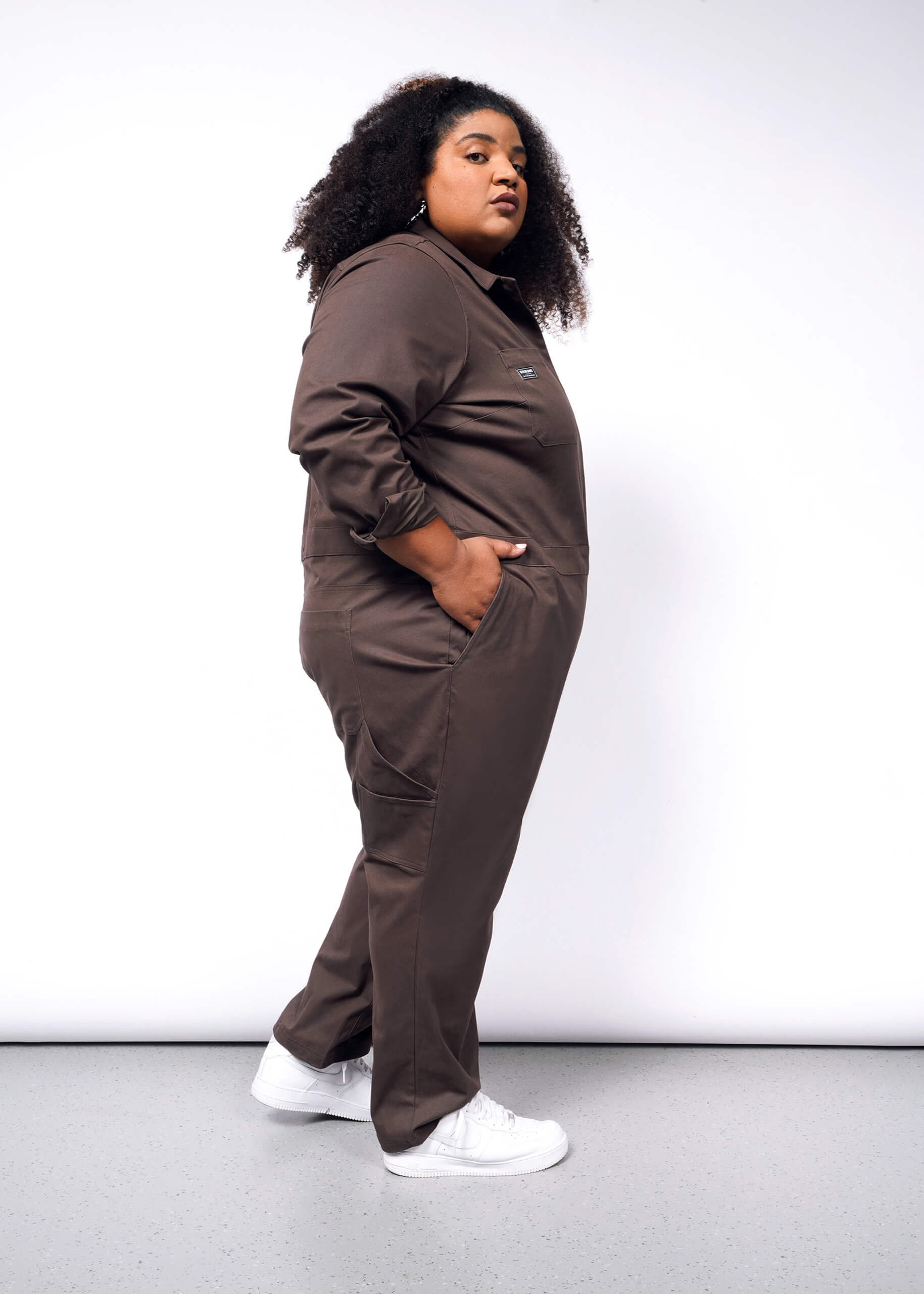 A person with curly hair stands sideways, wearing The Essential Long Sleeve Coverall in an oversized fit, made of brushed stretch cotton. Theyre sporting white sneakers and have one hand in their pocket, exuding confidence against a plain white background.